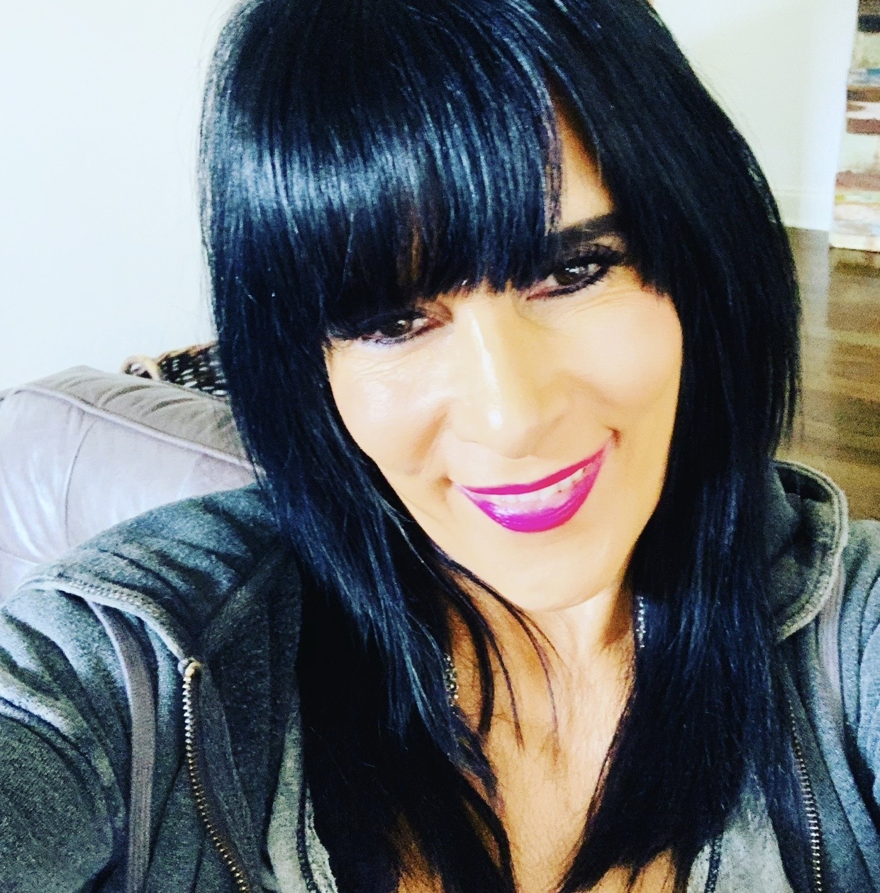 Woman with black hair and bangs smiling, wearing a dark jacket, in a room with a couch and wooden floor.