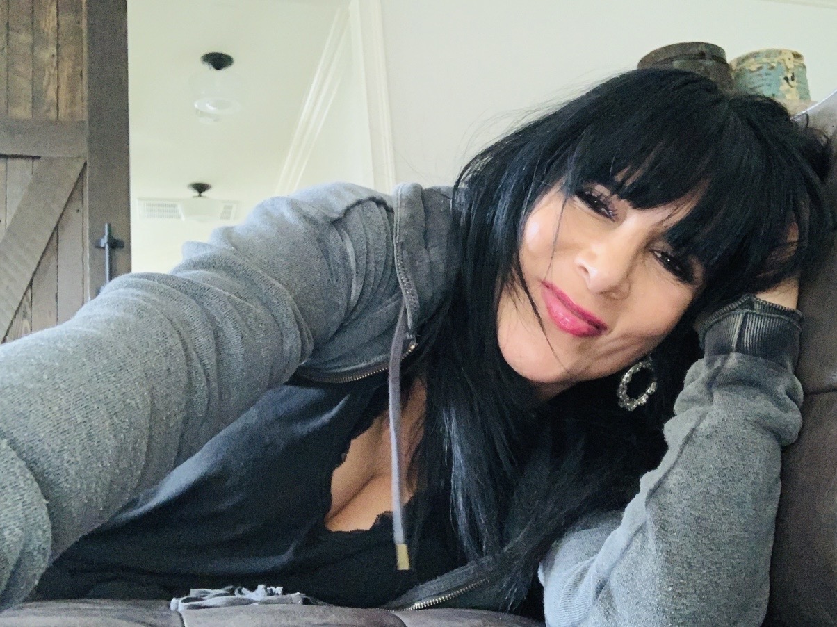 Woman with black hair and pink lipstick resting her head on her hand, smiling slightly, indoors.