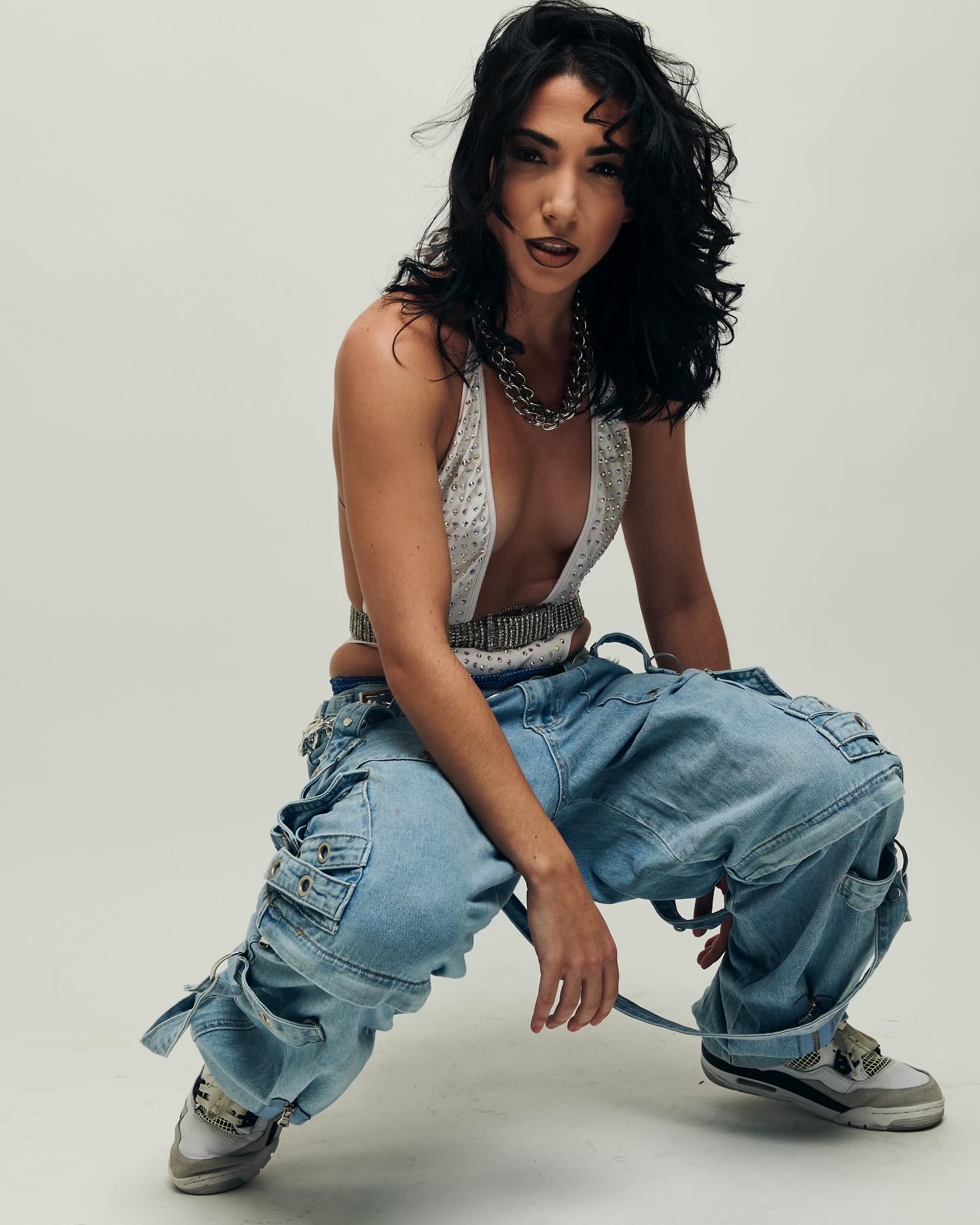 Woman with dark curly hair crouching, wearing a sleeveless top and baggy jeans, against a plain background.