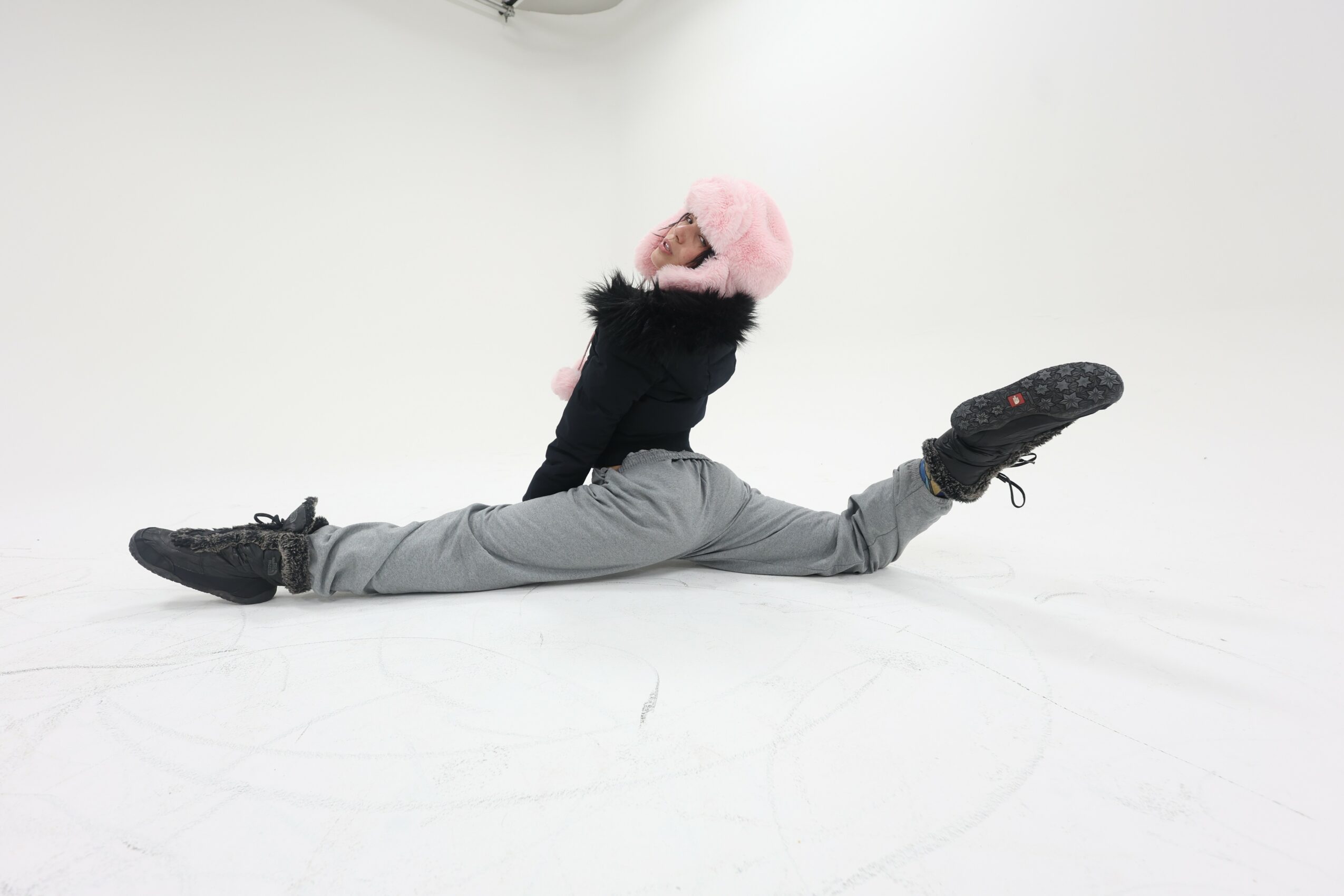 Person in winter clothing performing a split on the floor, wearing a pink hat and black gloves, against a white background.