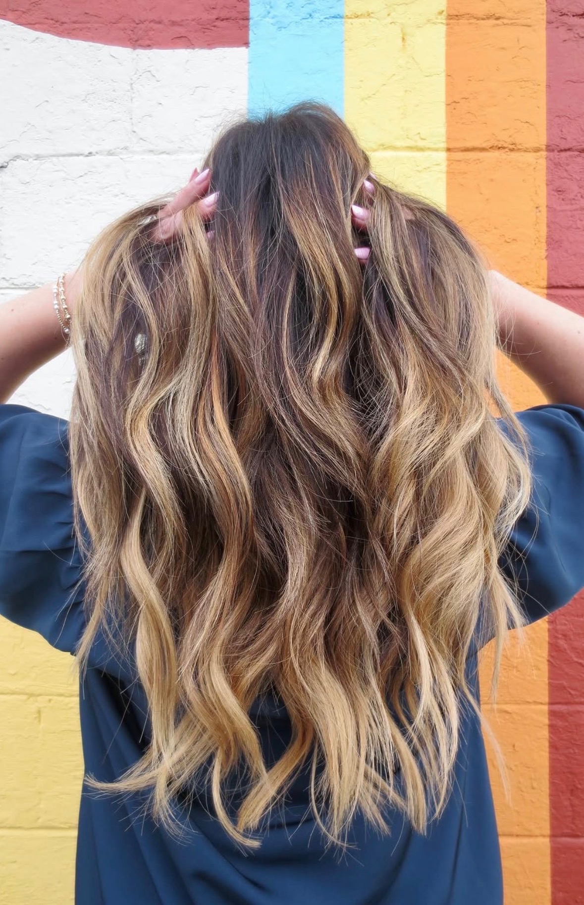 Person with long, wavy hair standing in front of a colorful wall, facing away, hands in hair, wearing a dark top.