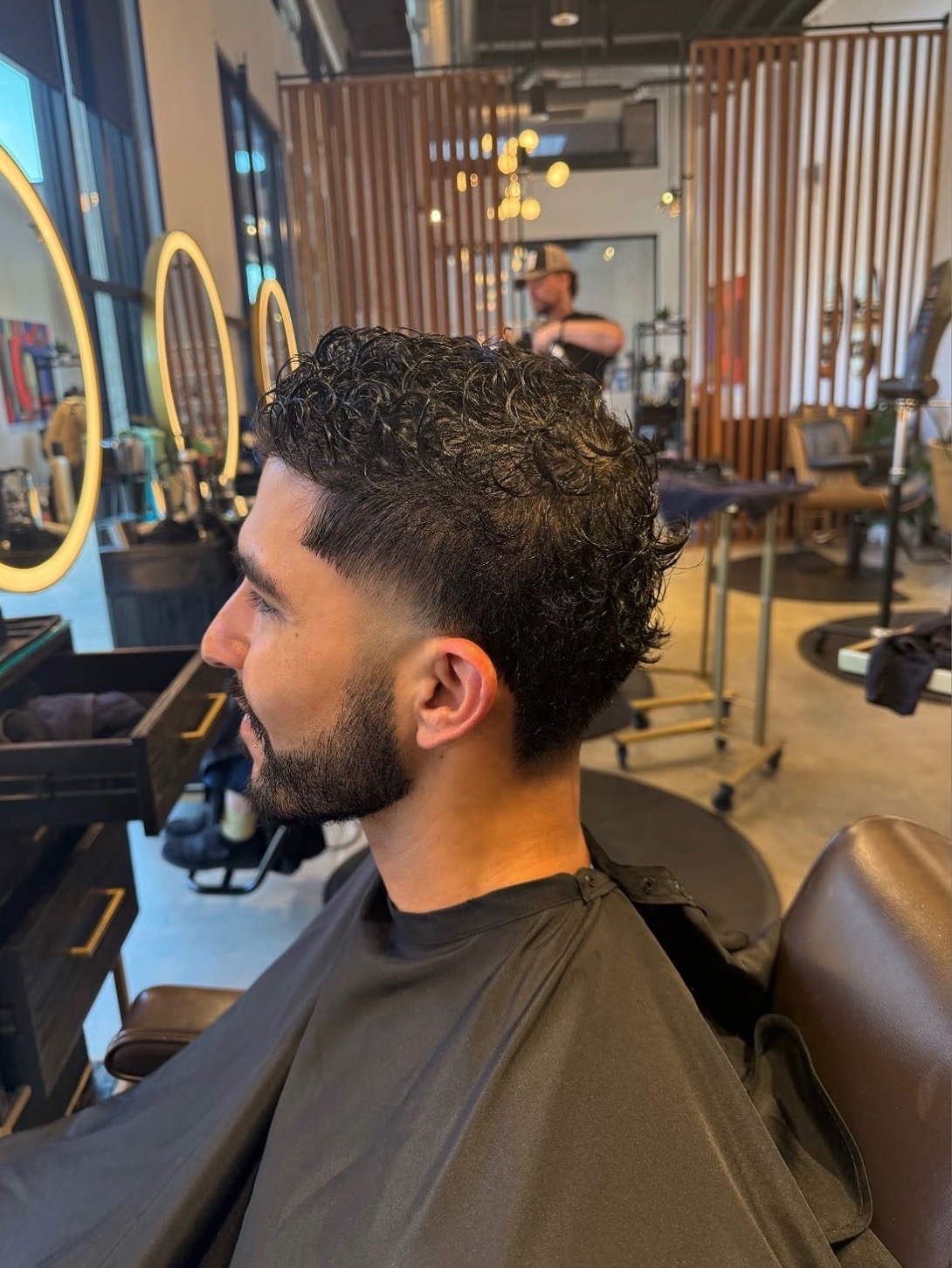 Man with curly hair and beard sitting in a barber shop chair, facing left, with mirrors and barber tools visible in background.