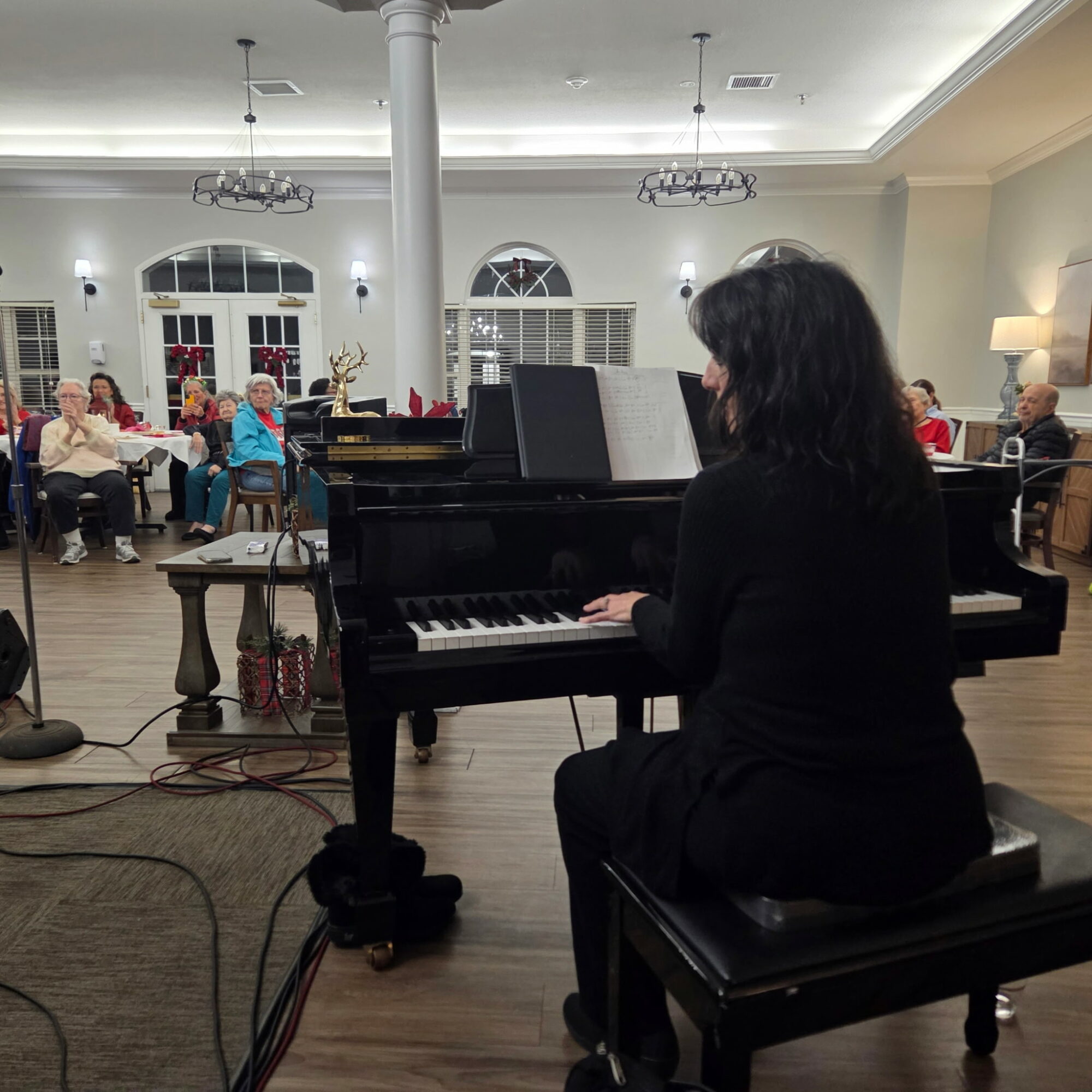 Person playing piano in a room with seated audience, large windows, and decorative lighting.