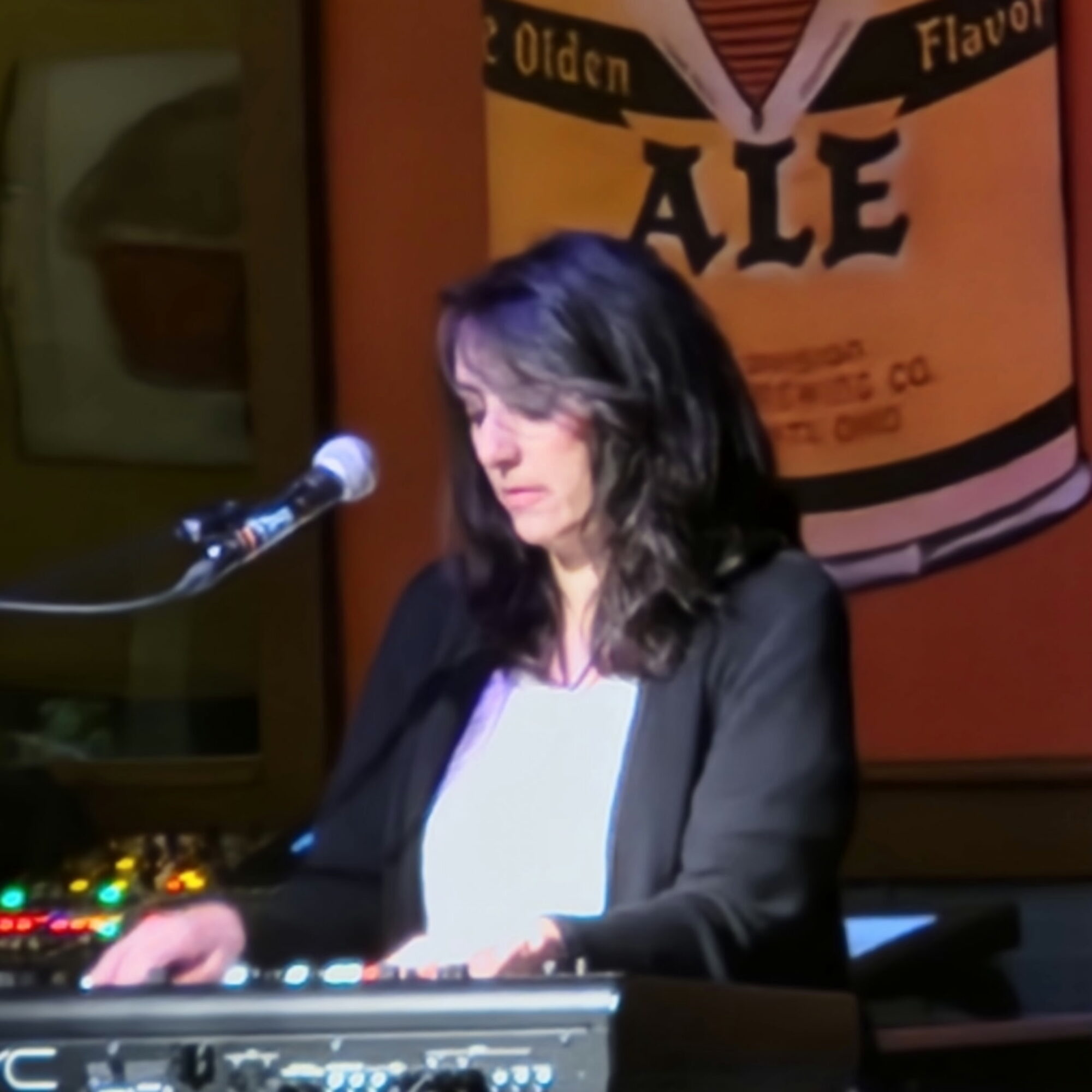 Woman with dark hair playing keyboard in front of a microphone, with a beer label poster in the background.