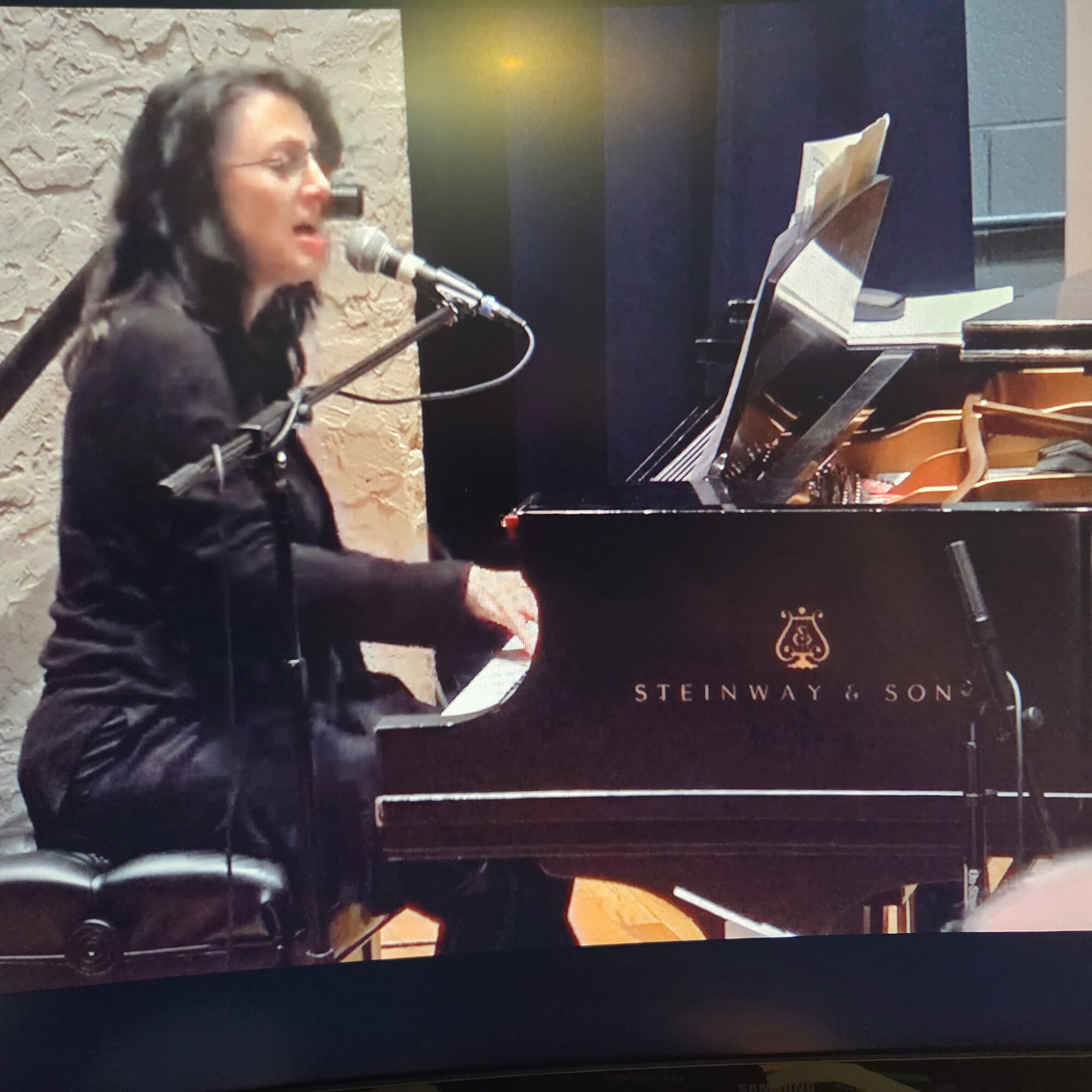 Woman singing into a microphone while playing a grand piano in a music studio, with sheet music on the piano.