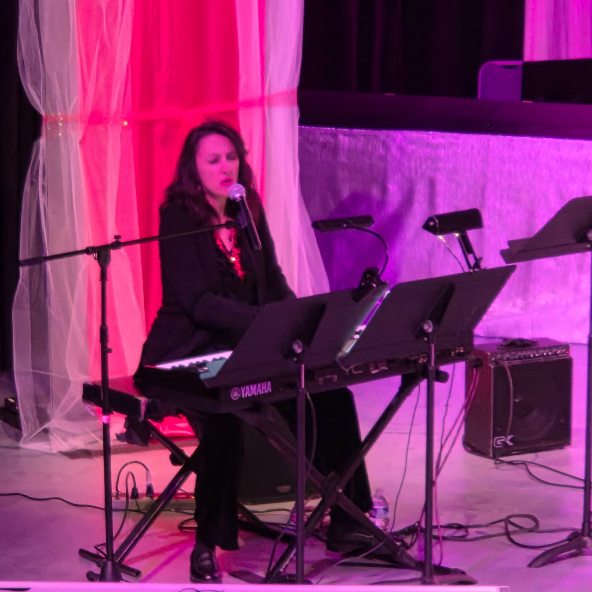 Woman singing and playing keyboard on stage with pink and purple curtains behind her.