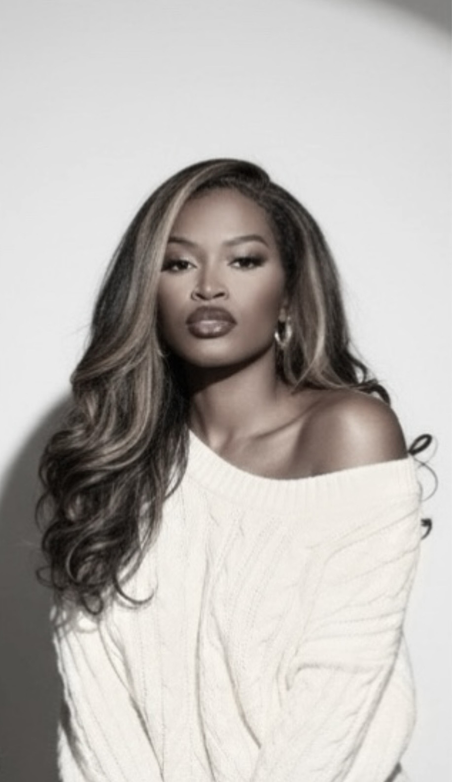 Woman with long wavy hair wearing a white off-shoulder sweater, looking at the camera, neutral background.