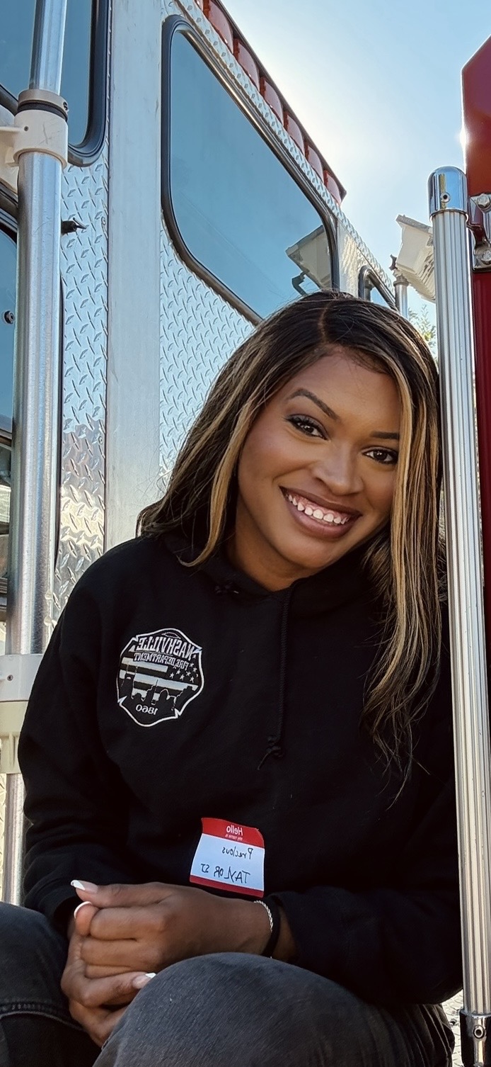 Smiling woman with long hair sitting outdoors near a large vehicle, wearing a black hoodie and a name tag.