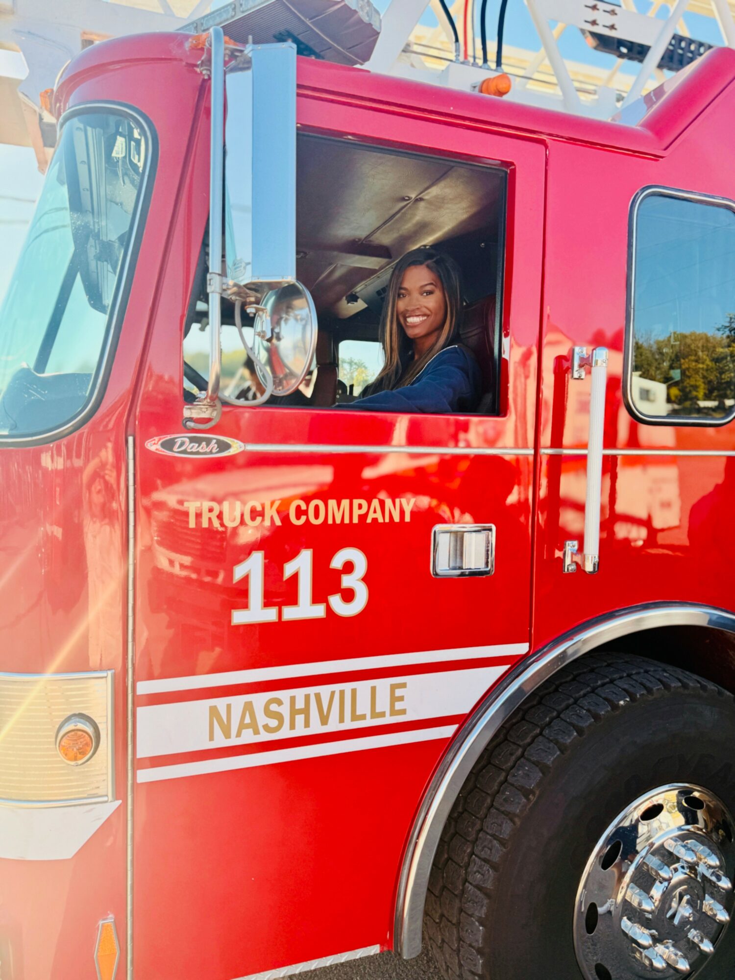 Woman smiling inside a red Nashville fire truck, number 113, with blue sky and trees in background.