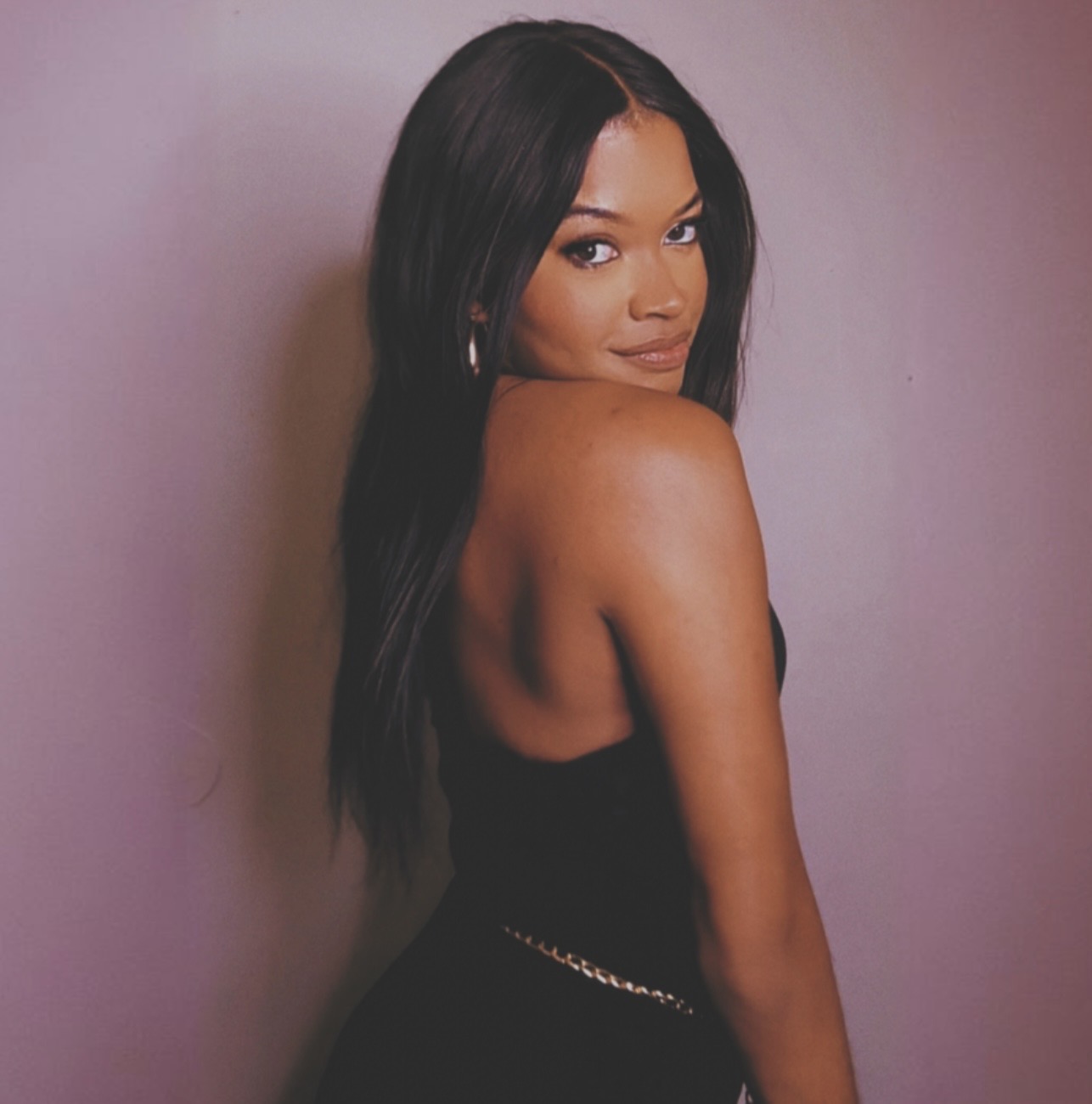 Young woman with long black hair looking over shoulder, wearing black top, standing against pink background.