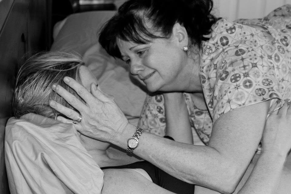 Woman gently touches a person's forehead, comforting them in bed, both smiling softly, in a grayscale photo.