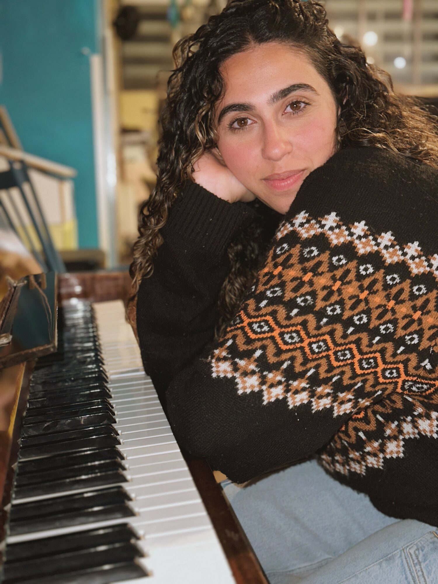 Young woman with curly hair resting her head on her hand, sitting near a piano, wearing a patterned sweater.