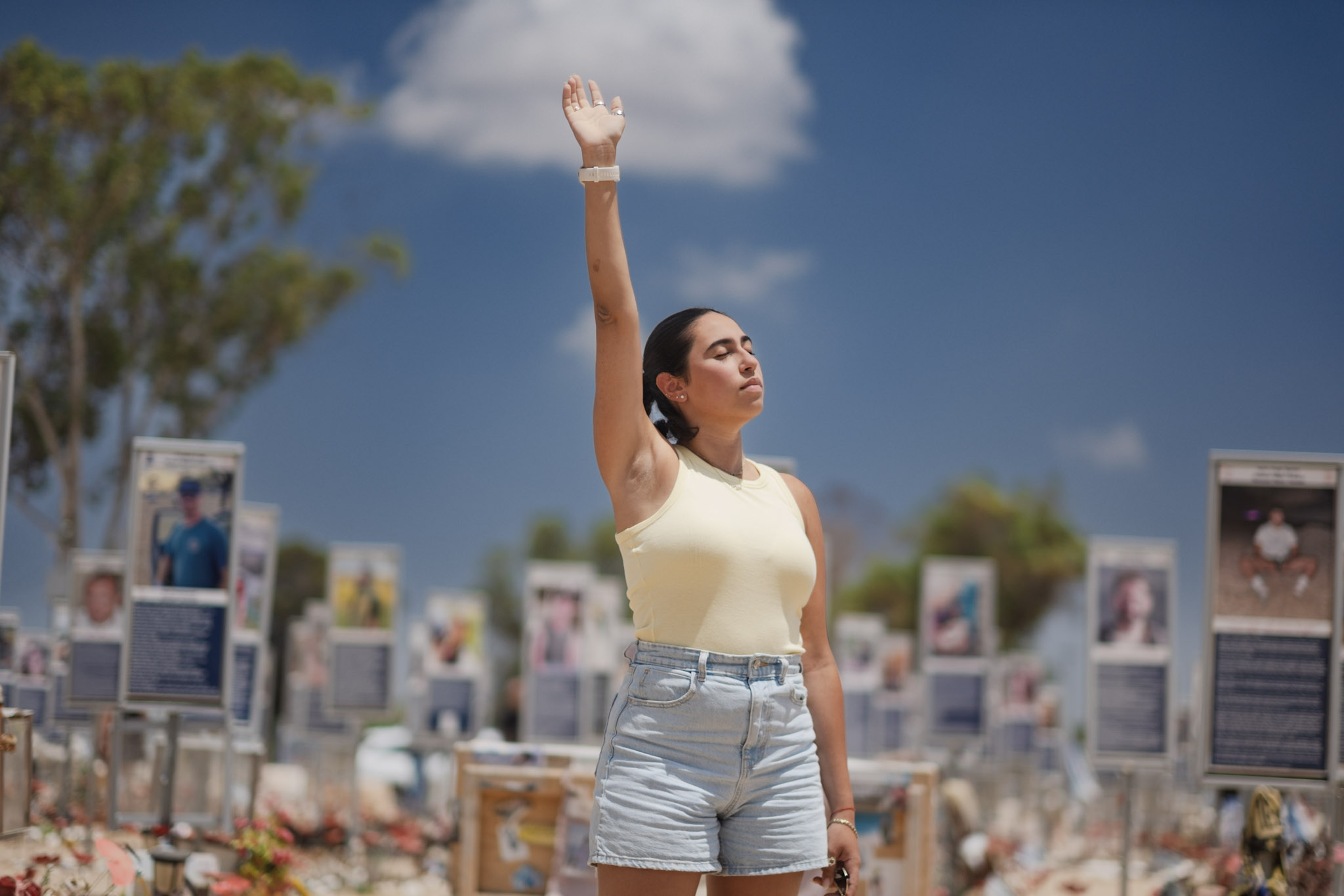 Woman in a white top and striped shorts raising her right arm outdoors with informational signs in the background.