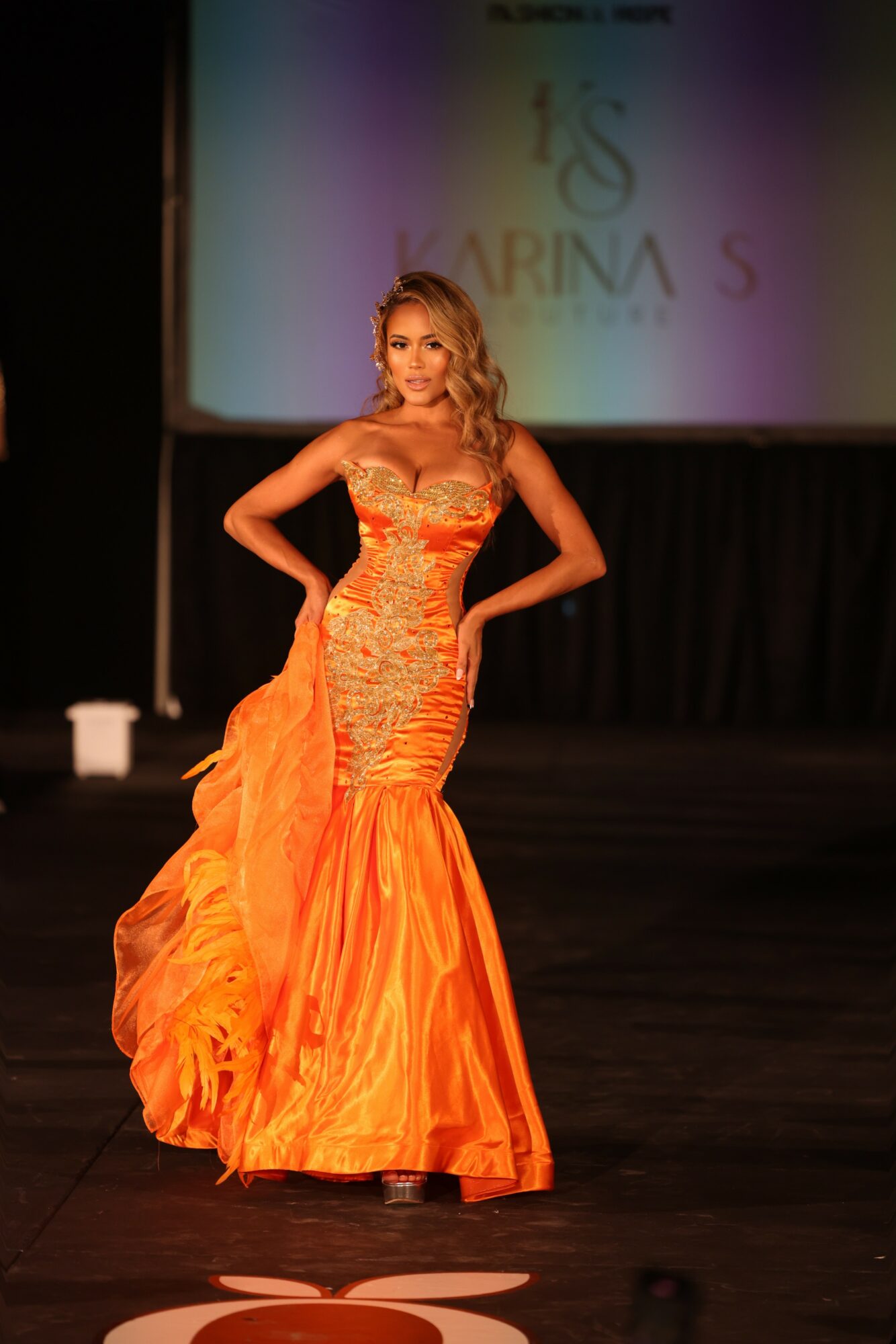 Woman in an orange gown on stage with a screen behind her displaying text and logo.