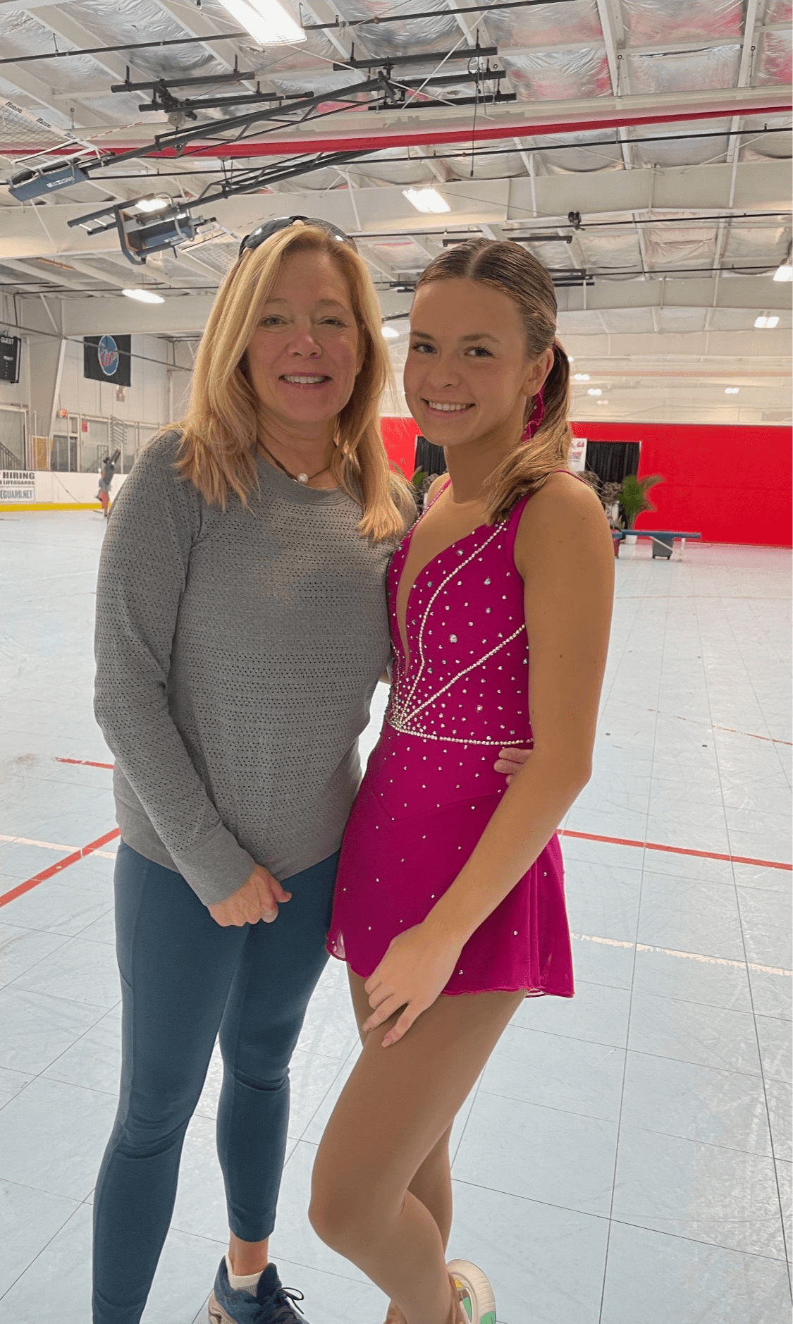 Two women standing together in an ice rink, smiling, with one wearing a gray sweater and the other in a pink figure skating dress.