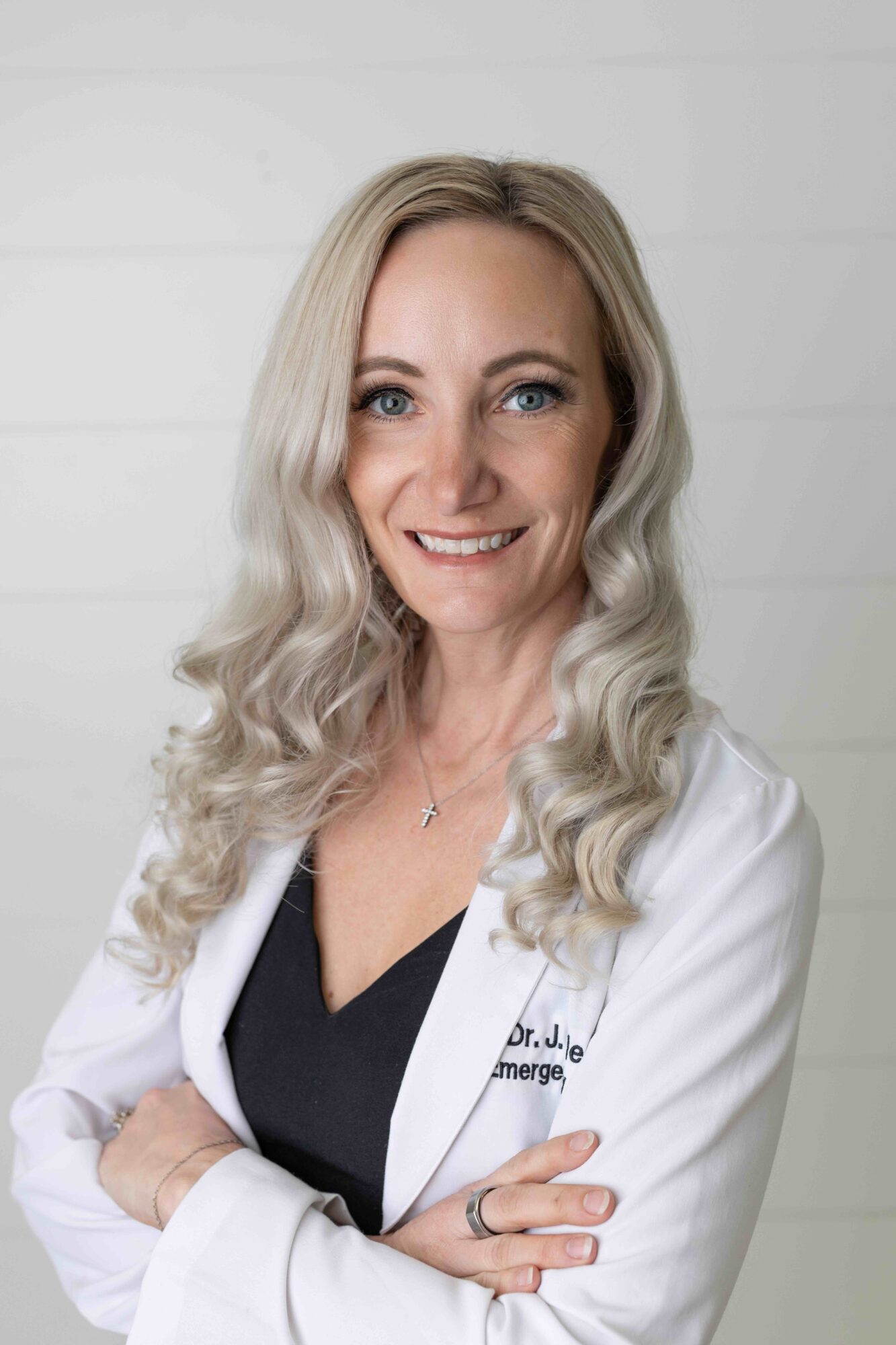 Woman with long wavy blonde hair smiling, wearing a white lab coat and black top, standing against a white background.