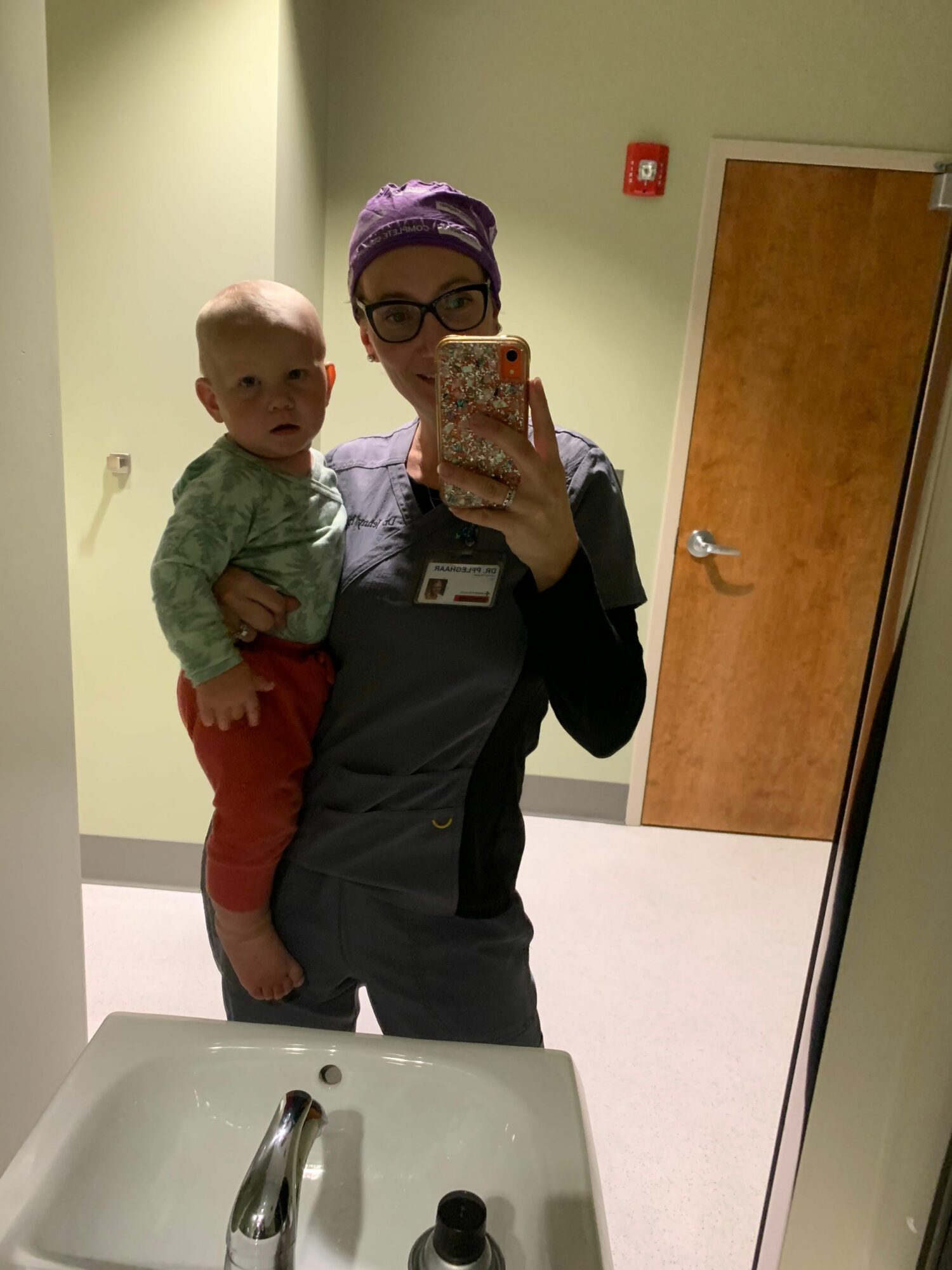 A person wearing glasses and a purple headband holds a child with light hair and green clothing in a room with a sink.