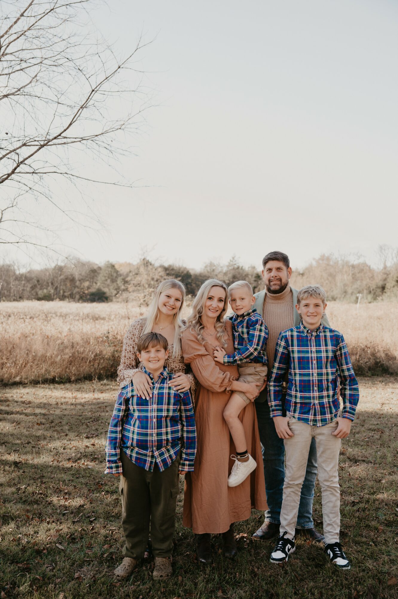 Family of five standing outdoors in a field with trees, smiling, with a clear sky background.