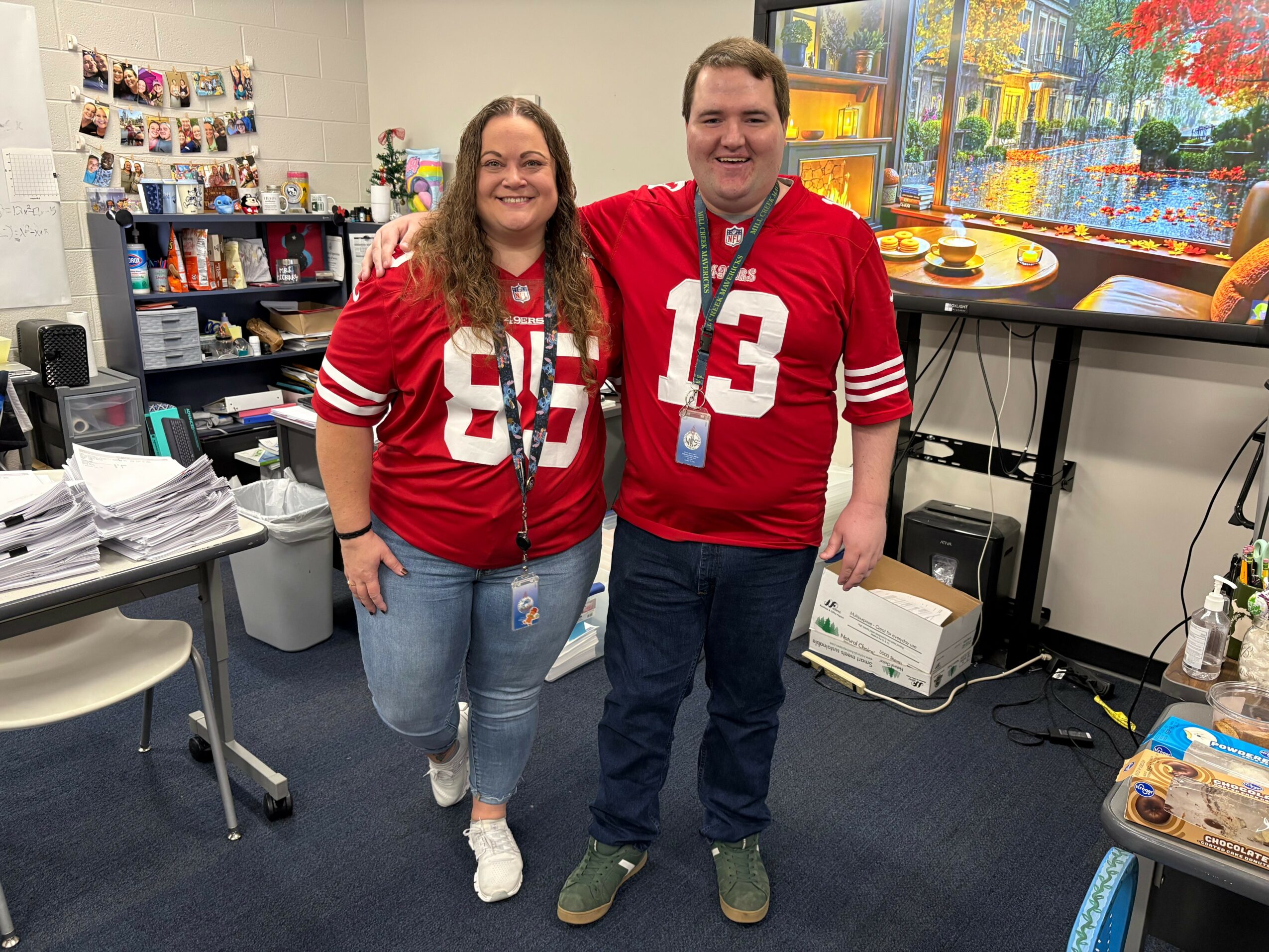Two people in red sports jerseys standing in an office, smiling, with a window showing trees outside.