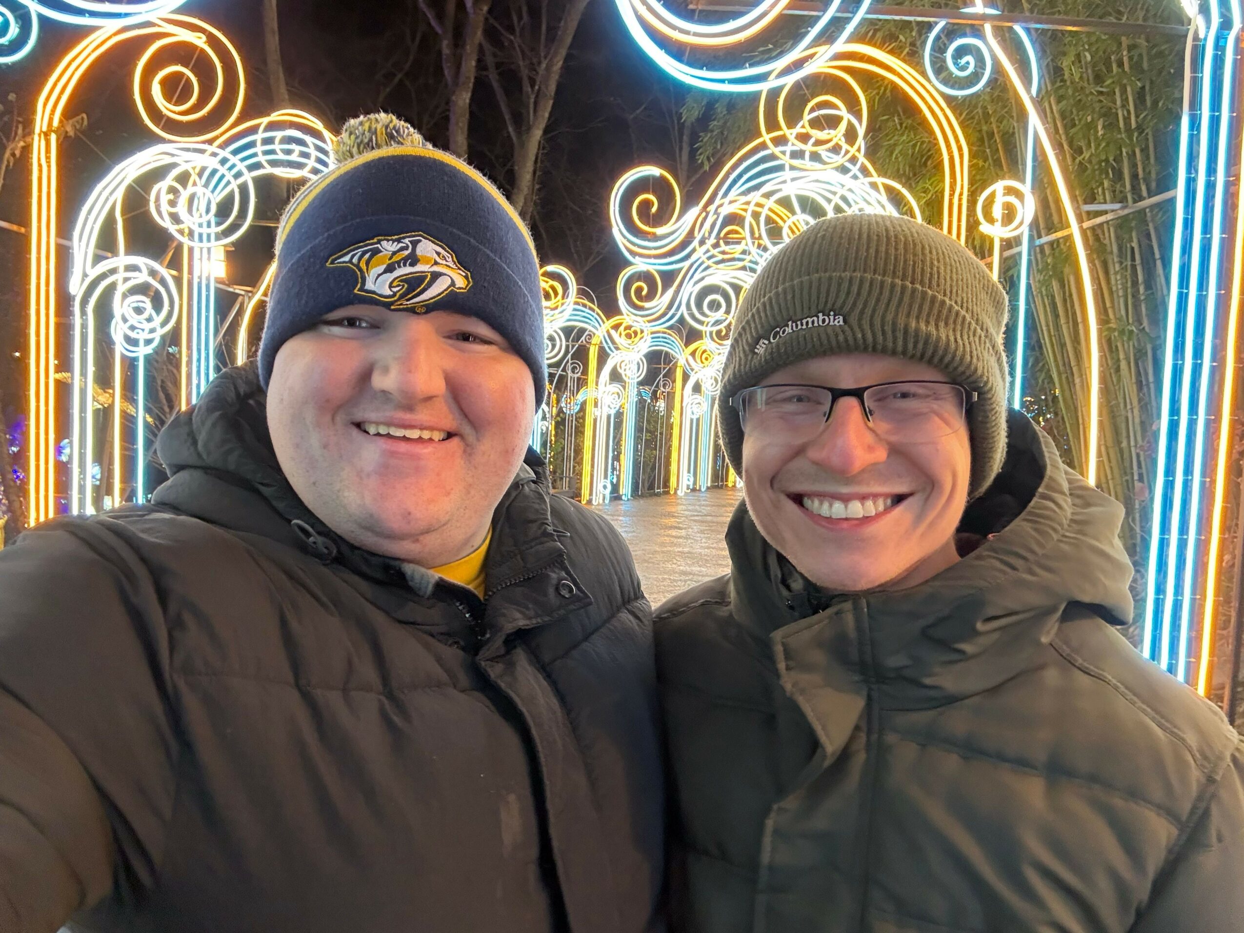 Two smiling people wearing winter clothing and hats, standing in front of colorful light decorations at night.