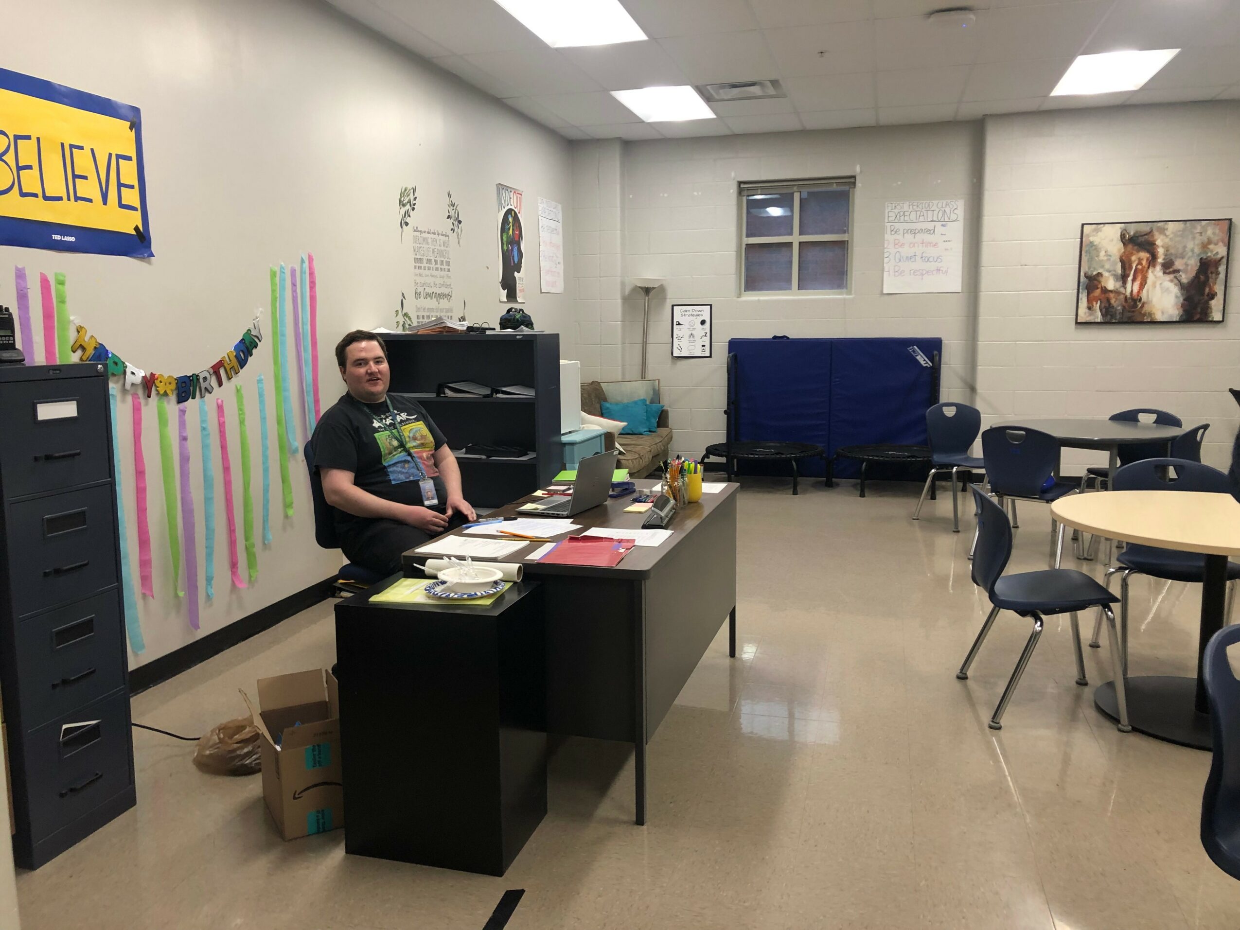 Person sitting at a desk in a classroom with colorful decorations and tables, white walls, and a window.