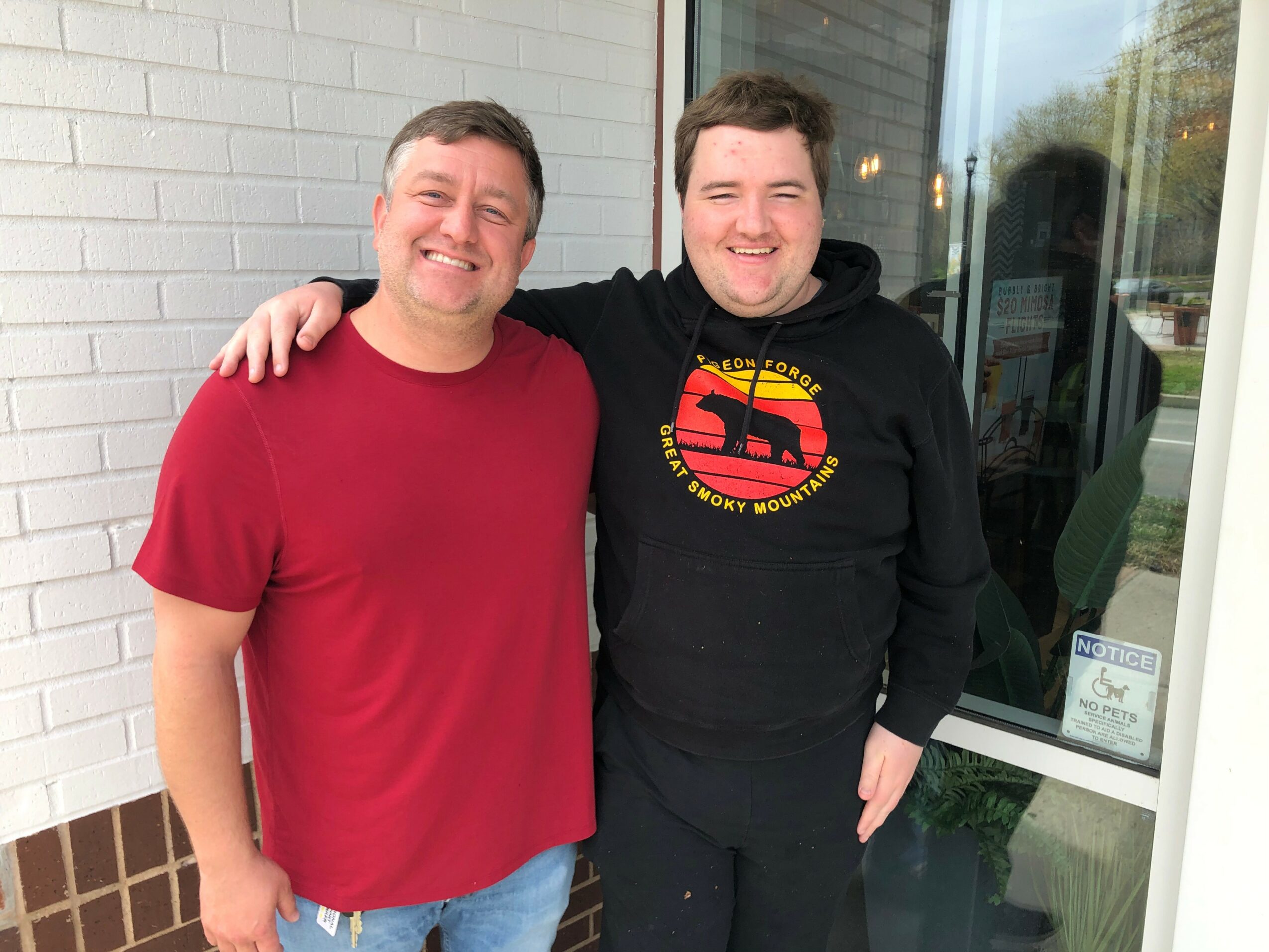 Two smiling men standing outside near a glass door, one with arm around the other, both casually dressed.