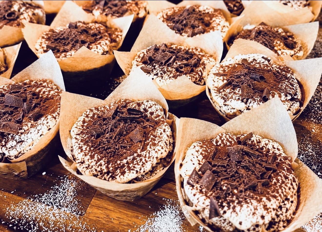 Multiple chocolate muffins in paper cups on a wooden surface, some dusted with powdered sugar.