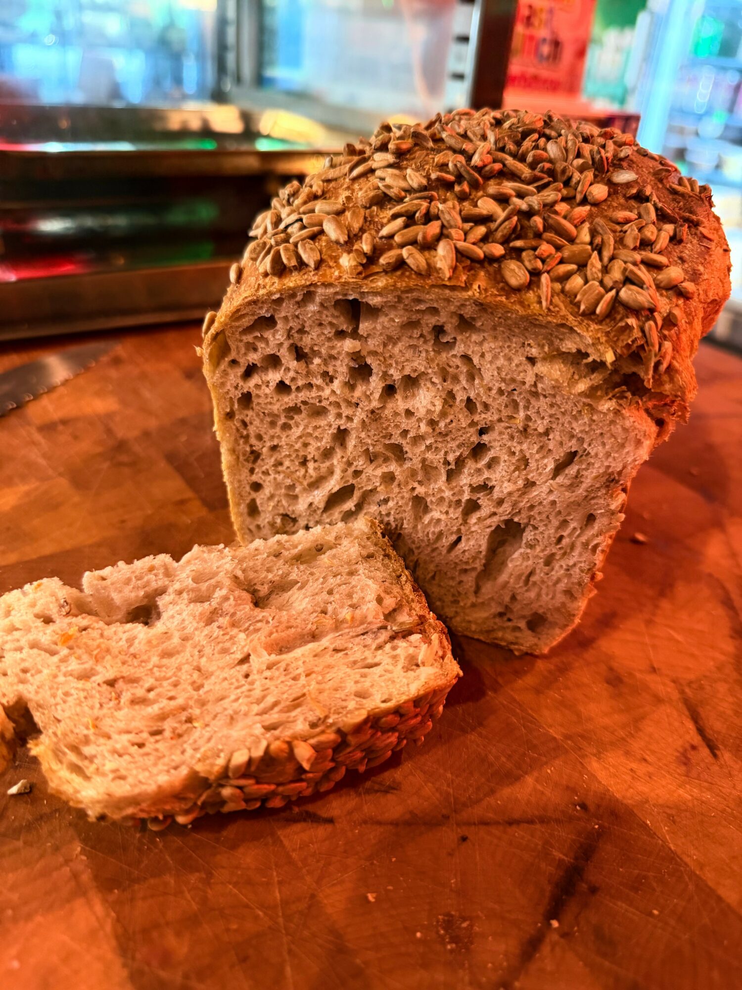 Loaf of bread with a slice cut, topped with sunflower seeds, on a wooden surface, colorful background, warm lighting.