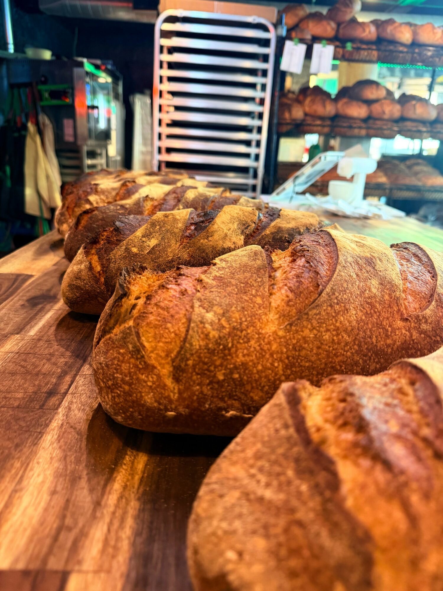 Several loaves of bread on a wooden surface in a bakery, with shelves of bread in the background.