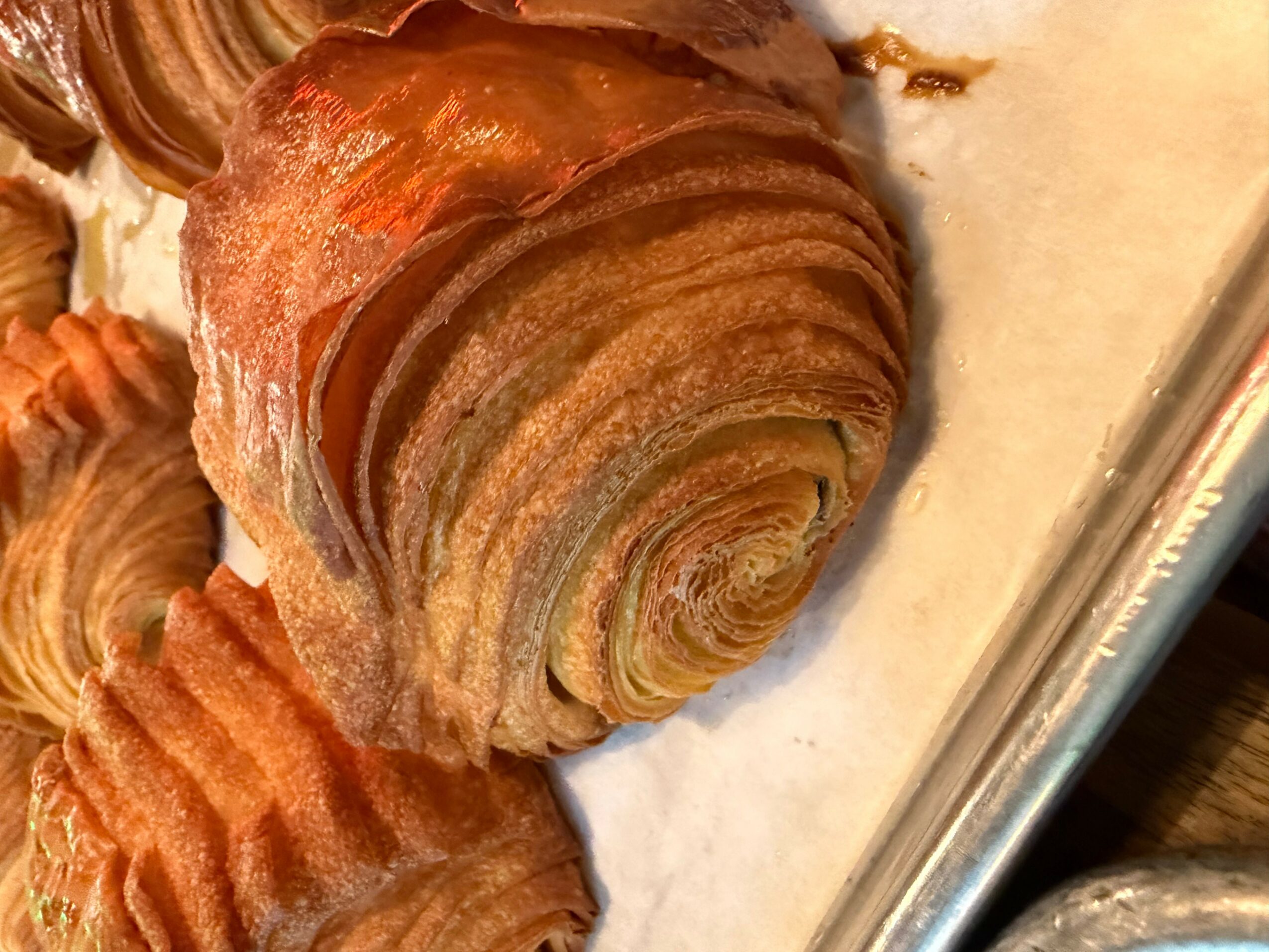 A close-up of a croissant on a baking sheet, showing its flaky, layered texture.