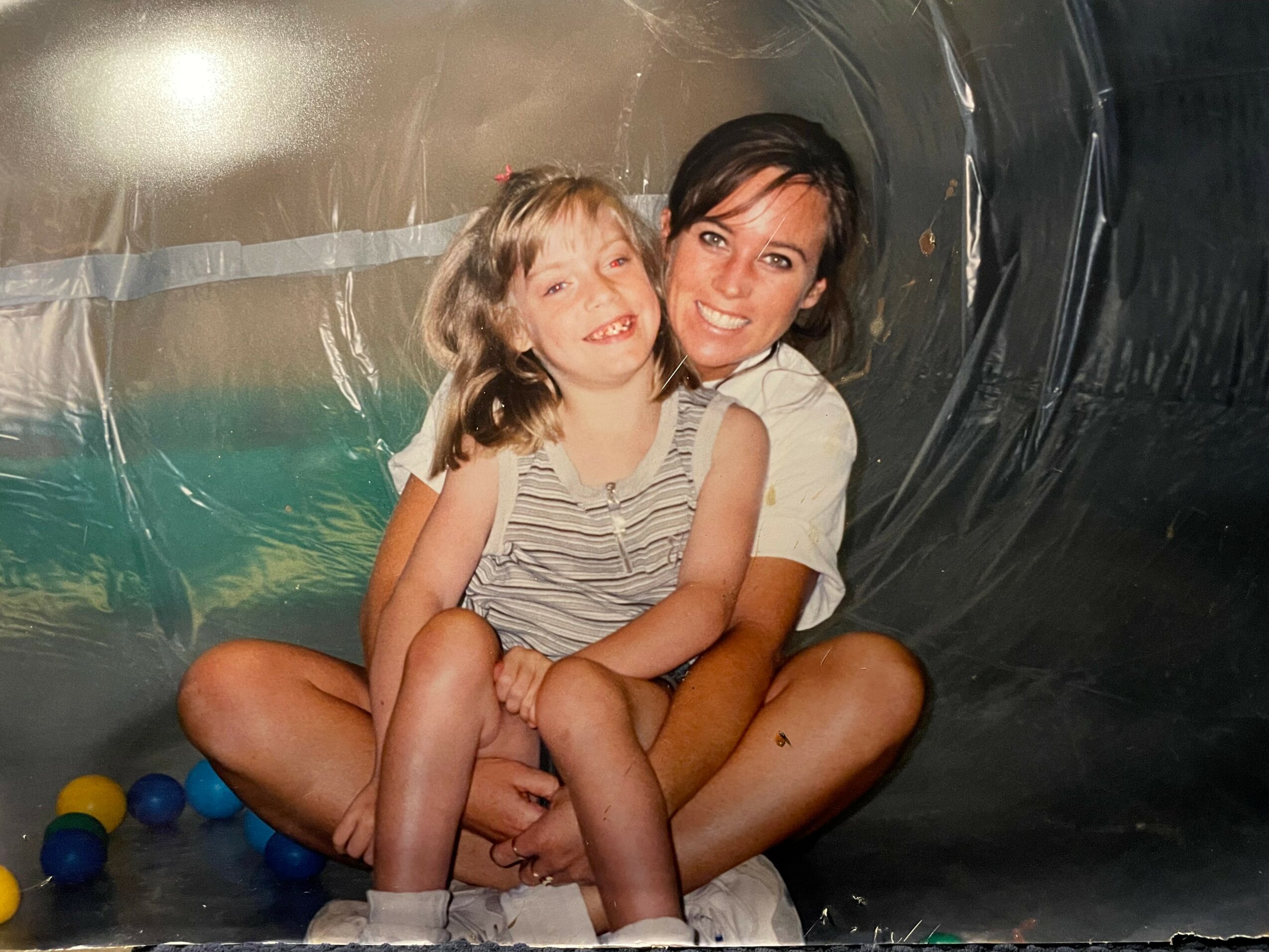 Two children sitting inside a metallic tunnel, smiling and hugging each other, with colorful balls around them.