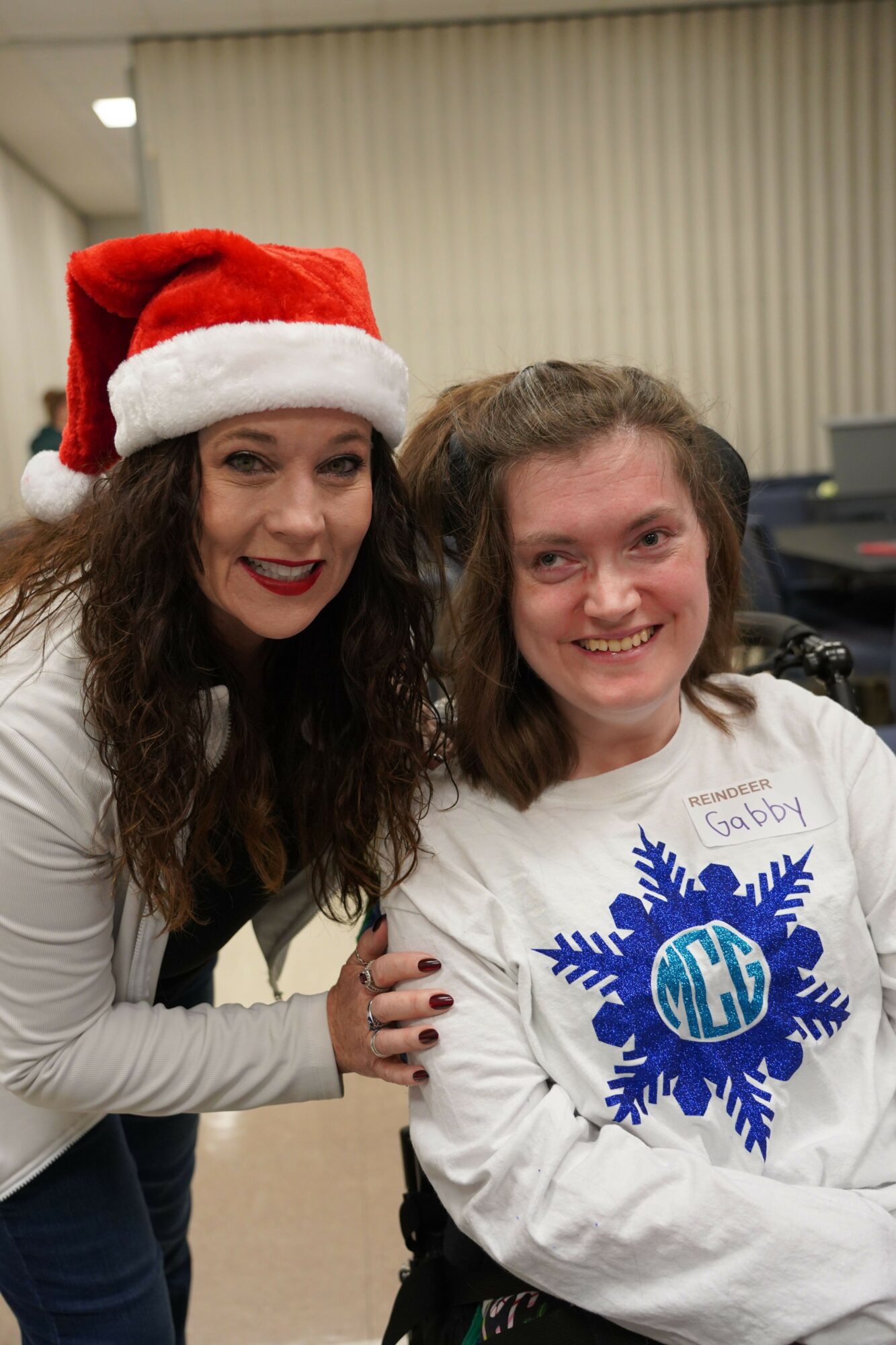 Two women smiling, one wearing a Santa hat, the other in a white sweatshirt with a blue snowflake design, indoors.