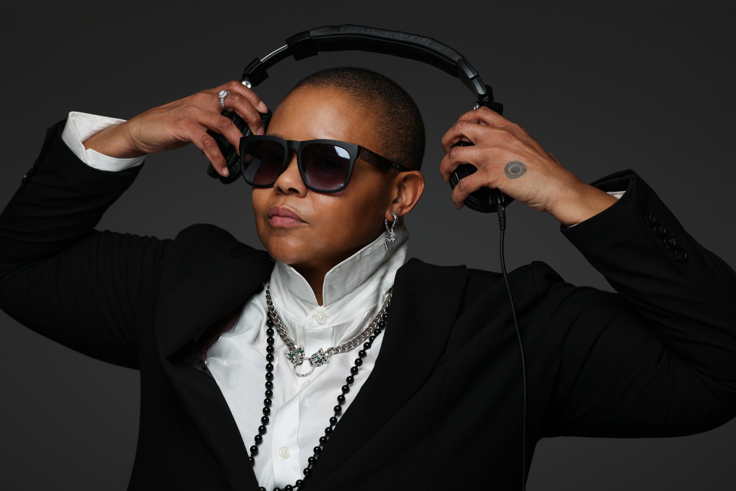 Person wearing sunglasses, headphones, and jewelry, holding headphones with both hands, against a dark background.