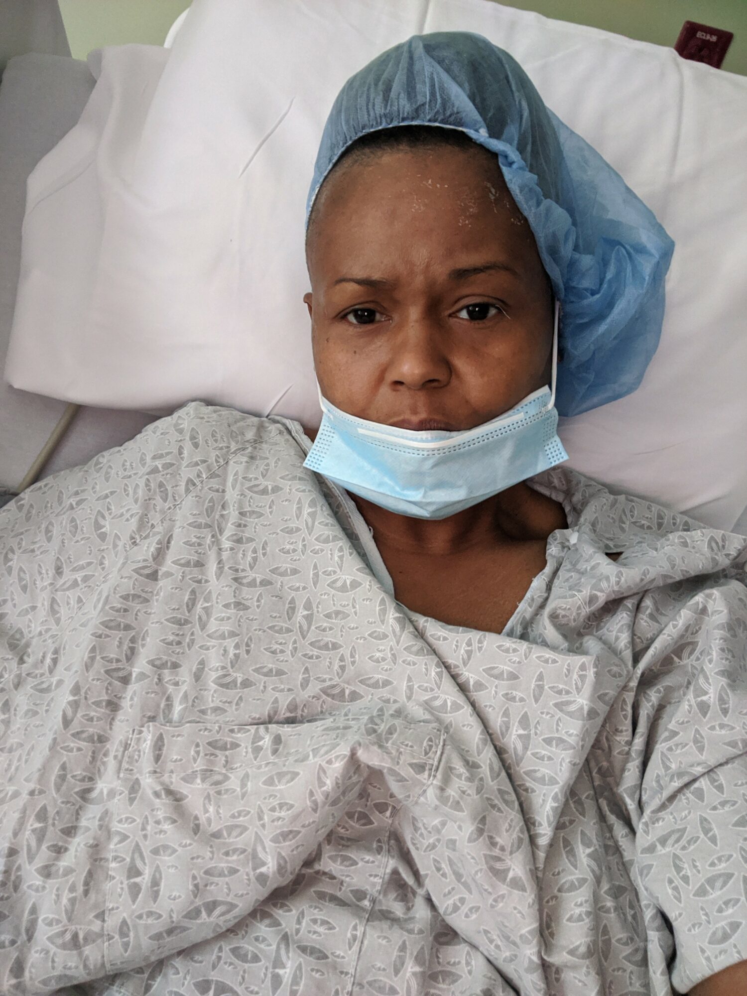 Woman in hospital bed wearing a blue surgical cap and face mask, looking at the camera.