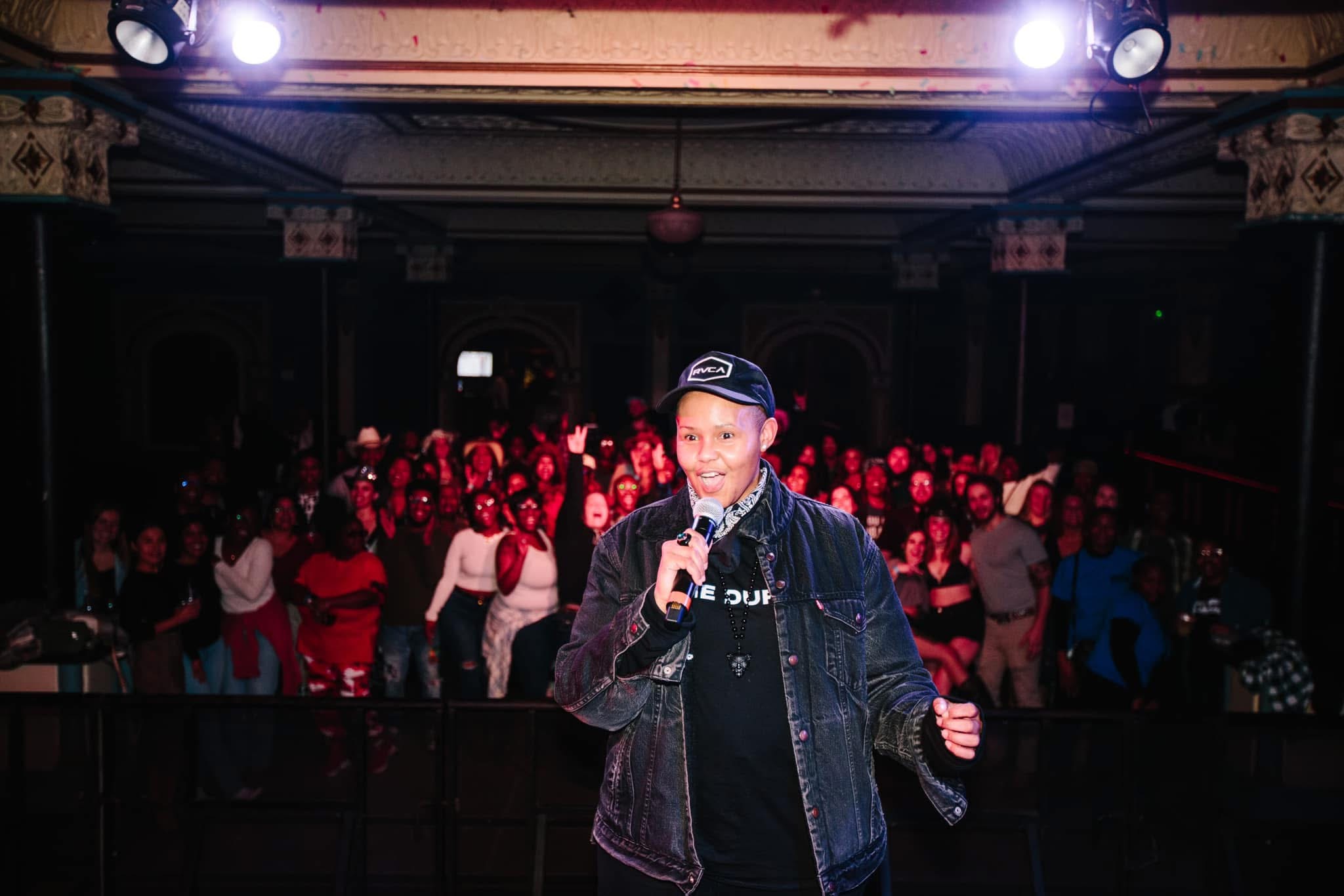 A young person holding a microphone on stage with a crowd in the background, stage lights overhead, in a dark venue.