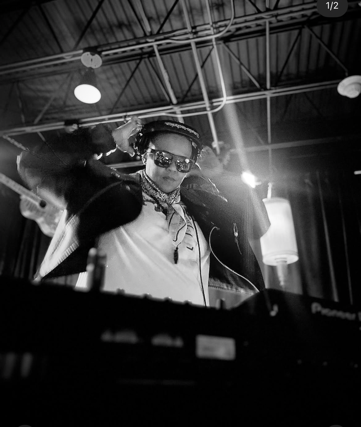 Person with sunglasses and braided hair, wearing a jacket, in a setting with industrial ceiling and lighting, possibly a DJ or performer.