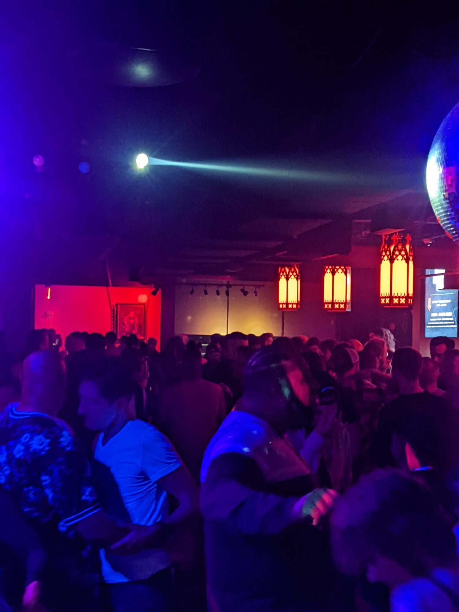 Crowd of people in a dark, colorful nightclub with bright lights and a large disco ball overhead.