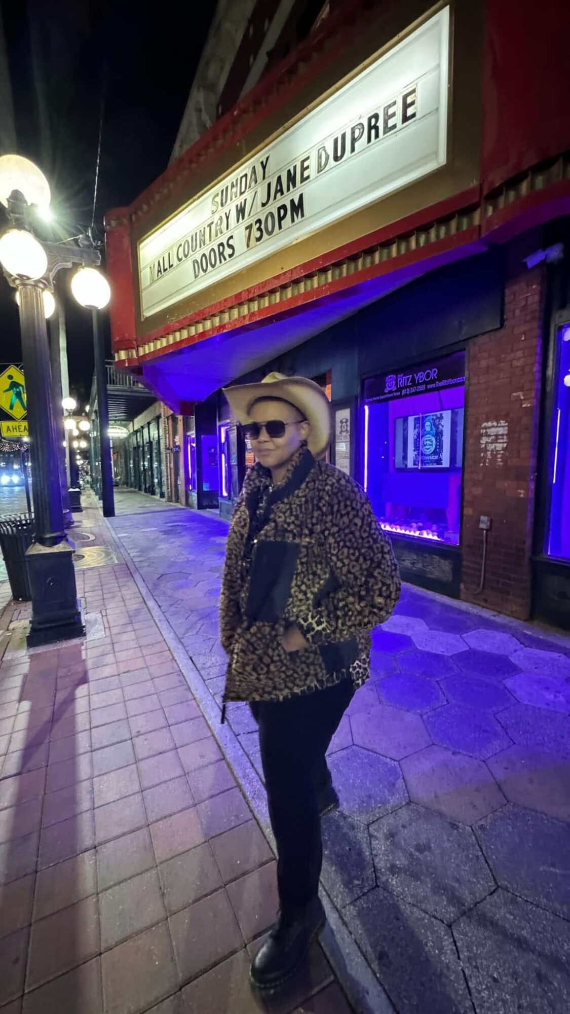 Person wearing sunglasses, hat, and patterned coat standing on sidewalk at night in front of theater marquee with purple lighting.