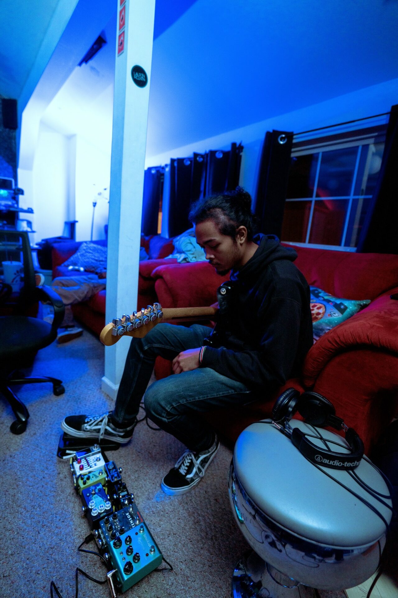 Person sitting on a red couch playing an electric guitar in a room with blue lighting, musical equipment nearby.