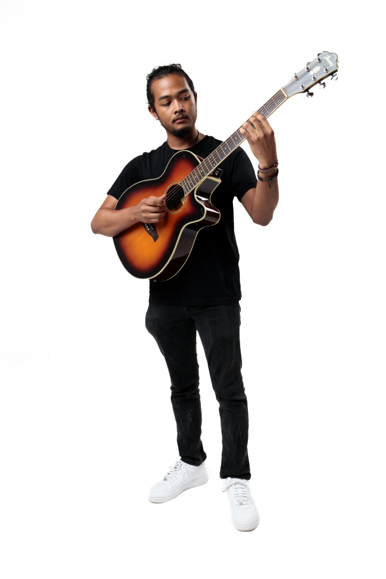 Person playing an acoustic guitar, standing against a white background, wearing a black shirt and pants, with white shoes.