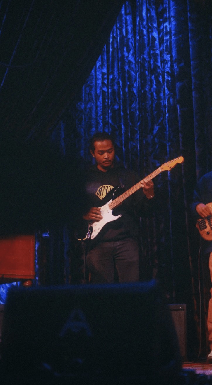 Person playing electric guitar on stage with blue curtain background, stage lighting, and another musician partially visible.
