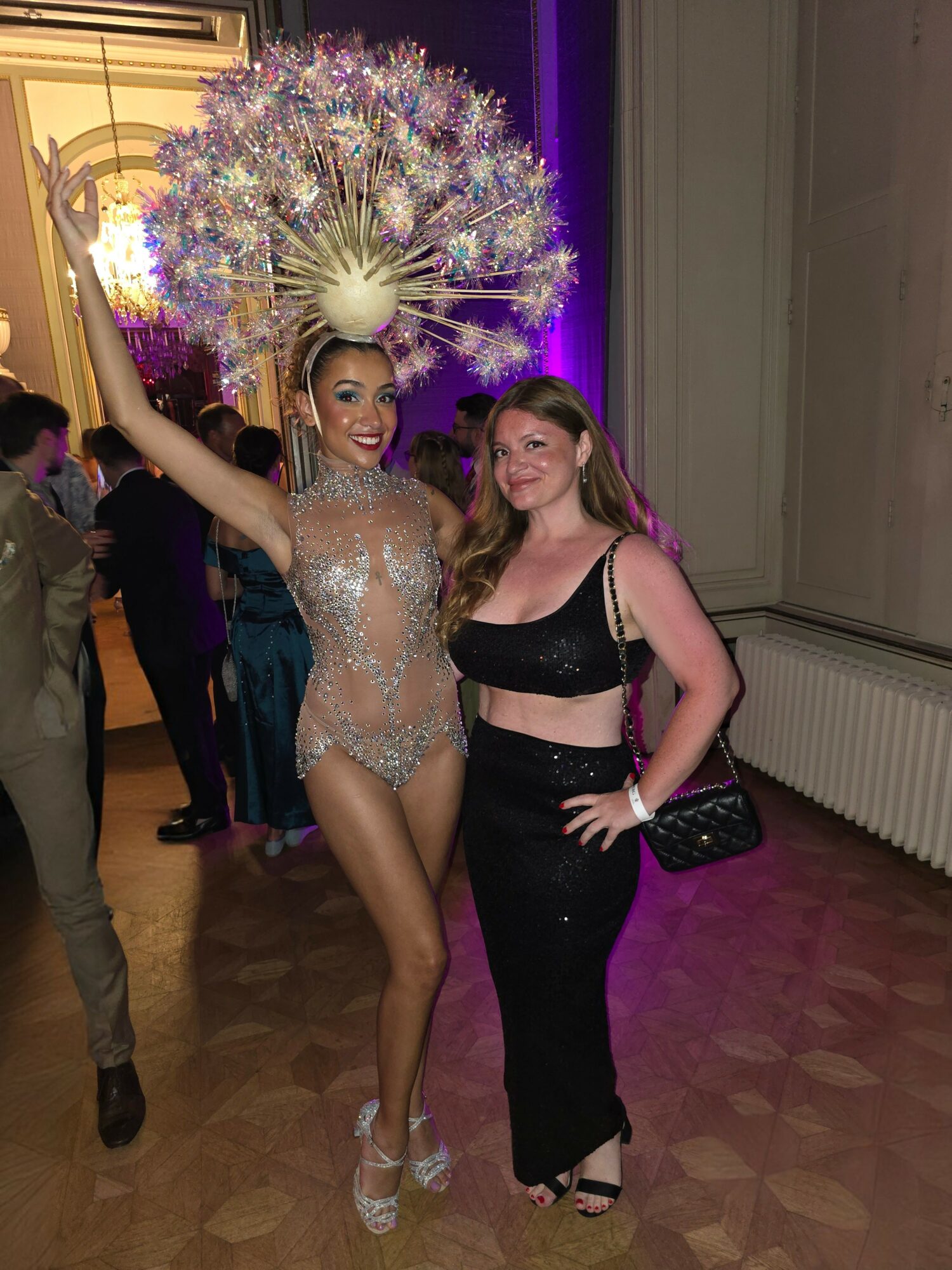 Two women pose together at an event, one in a sparkly outfit with a large feathered headpiece, the other in black attire.