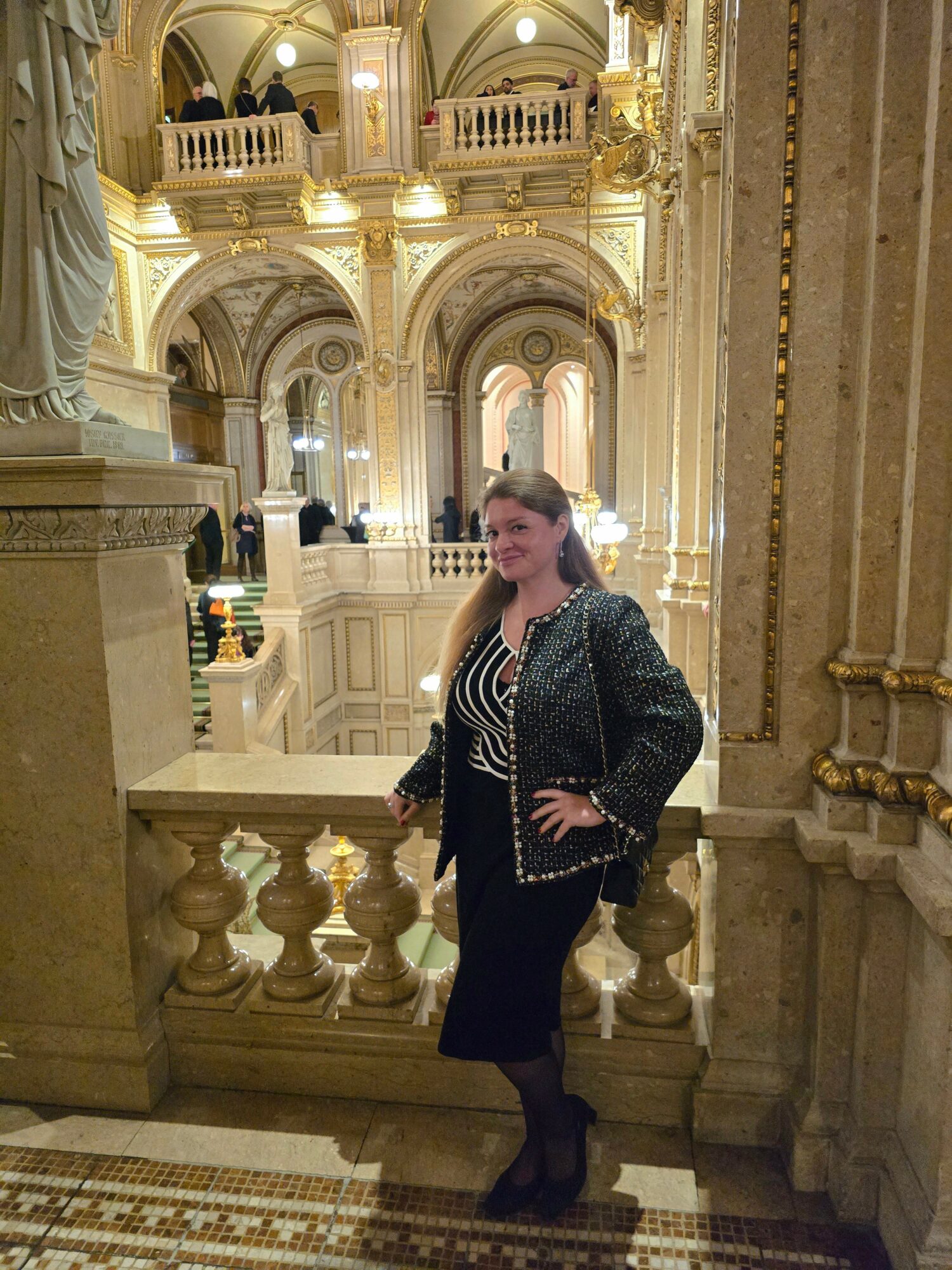 Woman standing in ornate, gold-lit interior with arches and statues, smiling, wearing a patterned top and dark skirt.