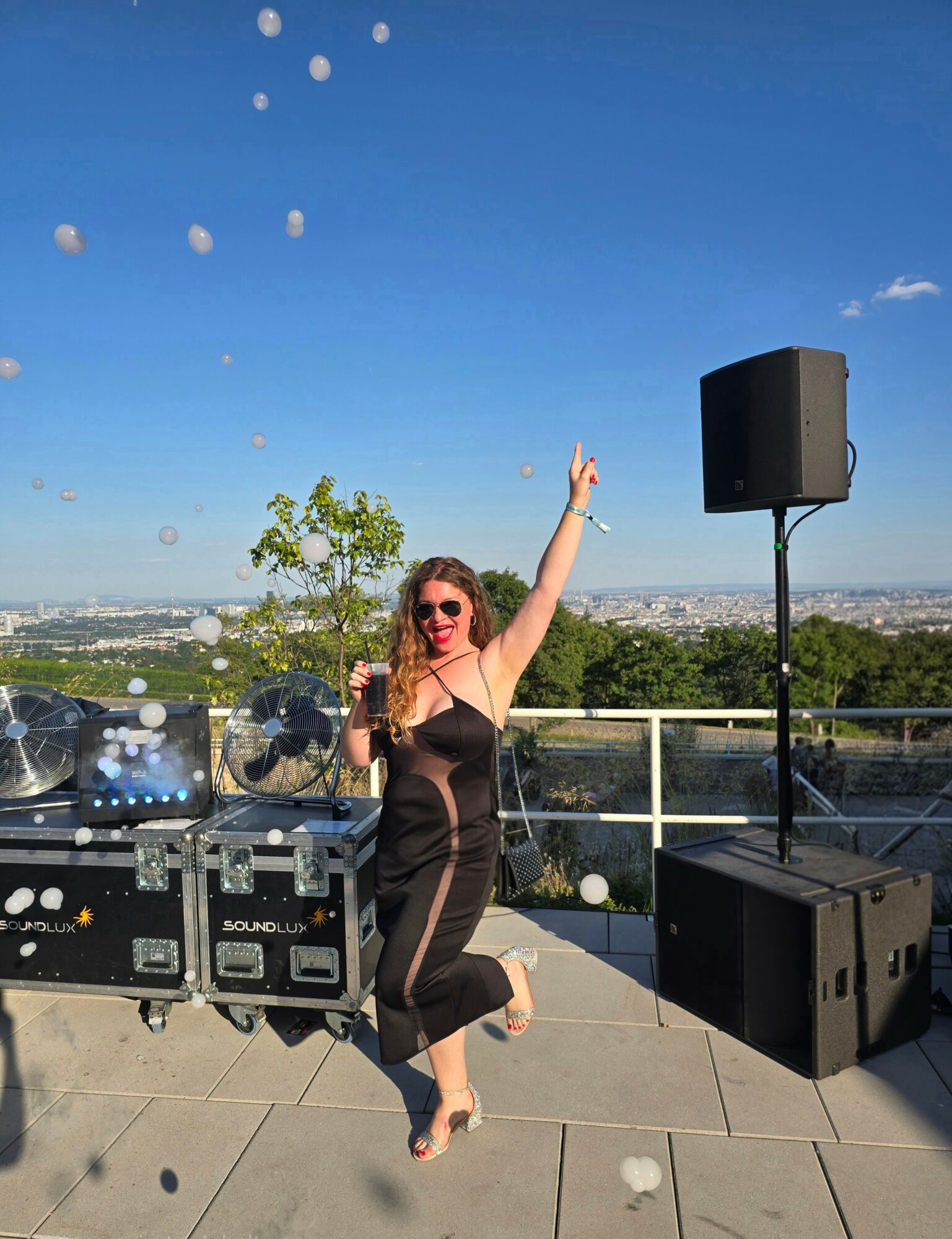 Woman in black dress and sunglasses dancing outdoors on a rooftop with DJ equipment and a speaker, cityscape in background.