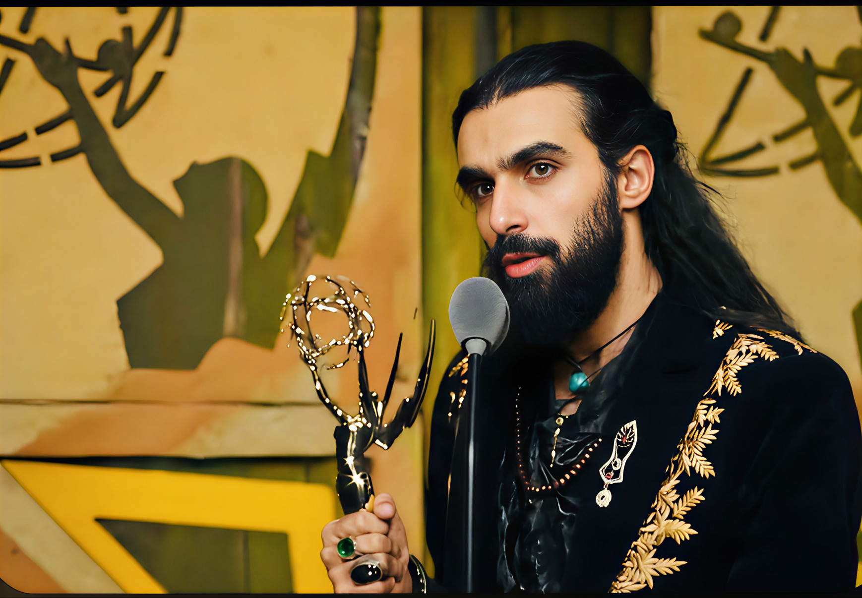 Man with long dark hair and beard holding an award, speaking into a microphone, with a colorful background.