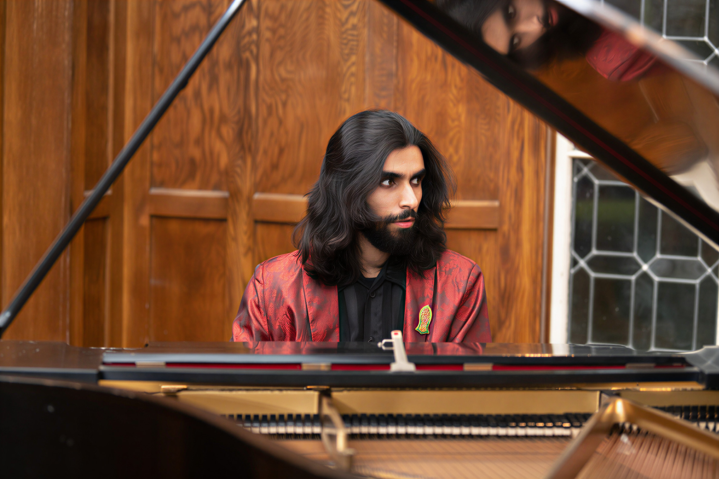 Young man with long dark hair and beard, wearing a red jacket, sitting at a piano, looking to the side.