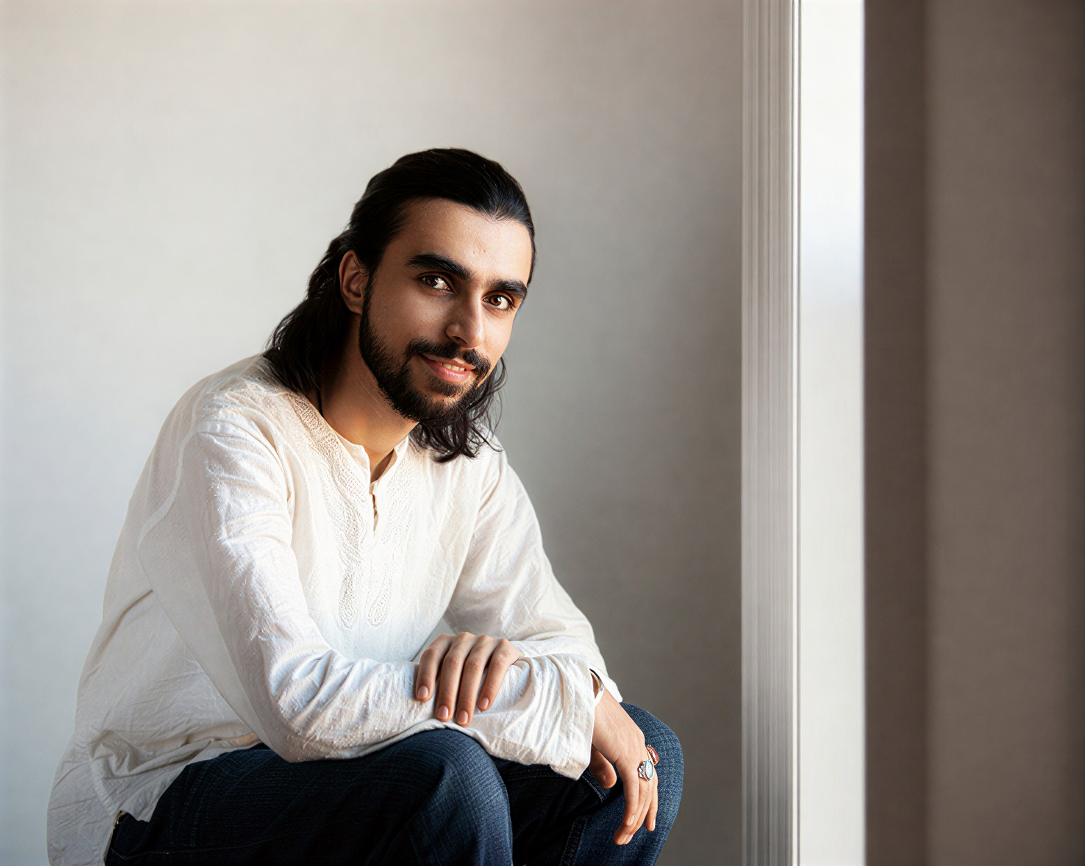 Person with long dark hair and beard sitting by a window, wearing a white shirt, looking at the camera.