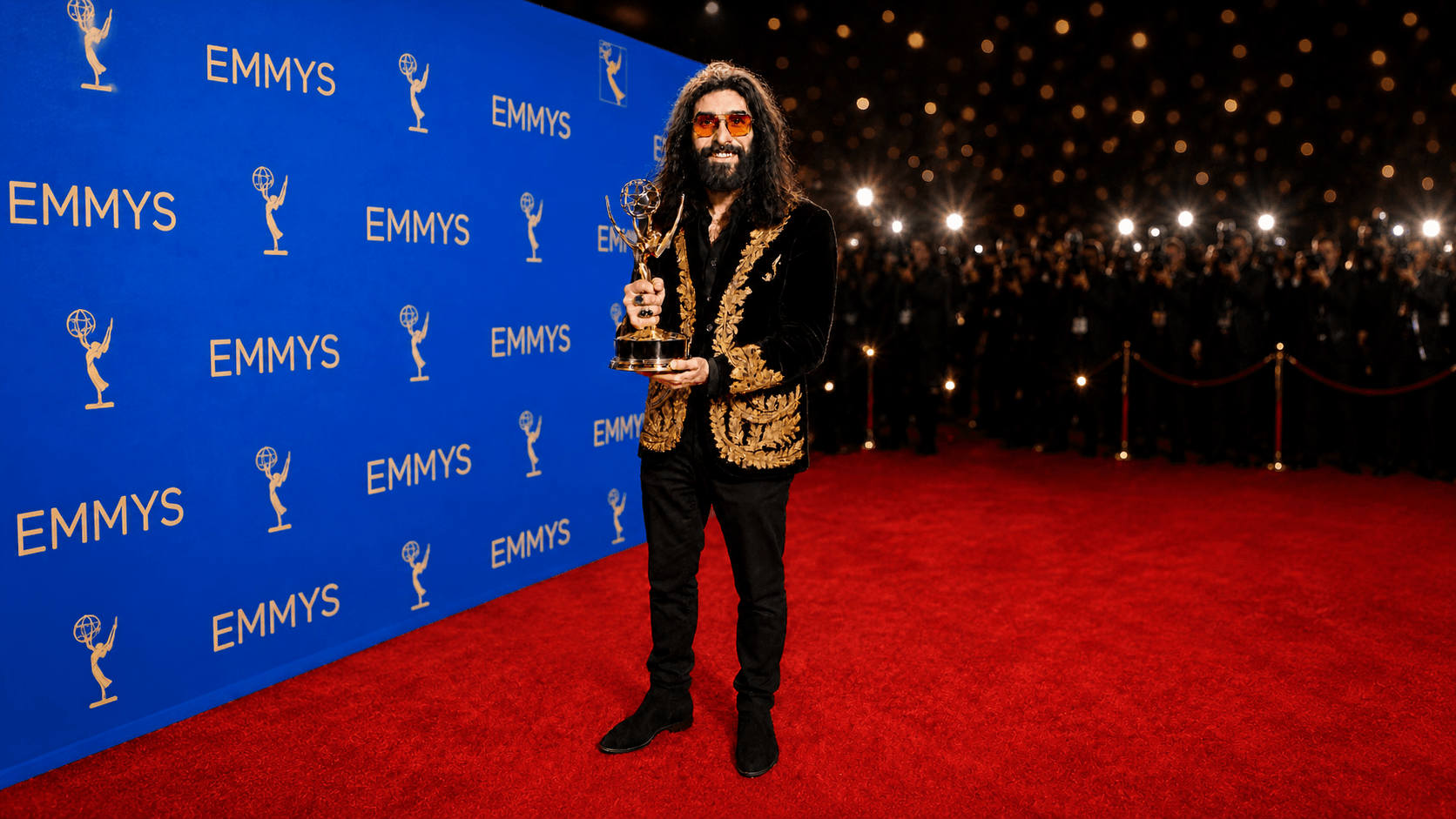 Person with long hair and beard holding an award on red carpet, backdrop with Emmy logos, and bright lights in background.