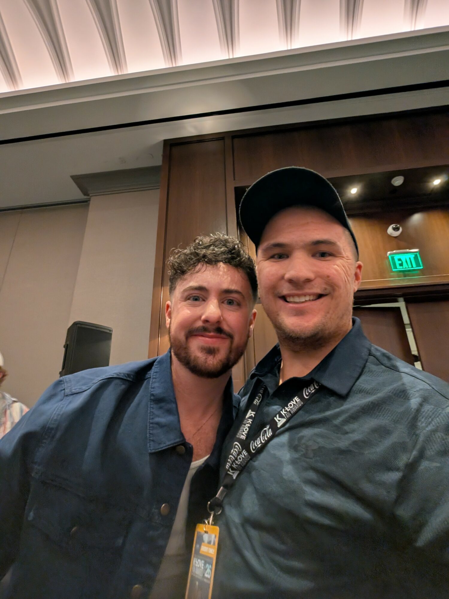 Two men smiling, one wearing a cap, in an indoor setting with wood paneling and an exit sign behind them.