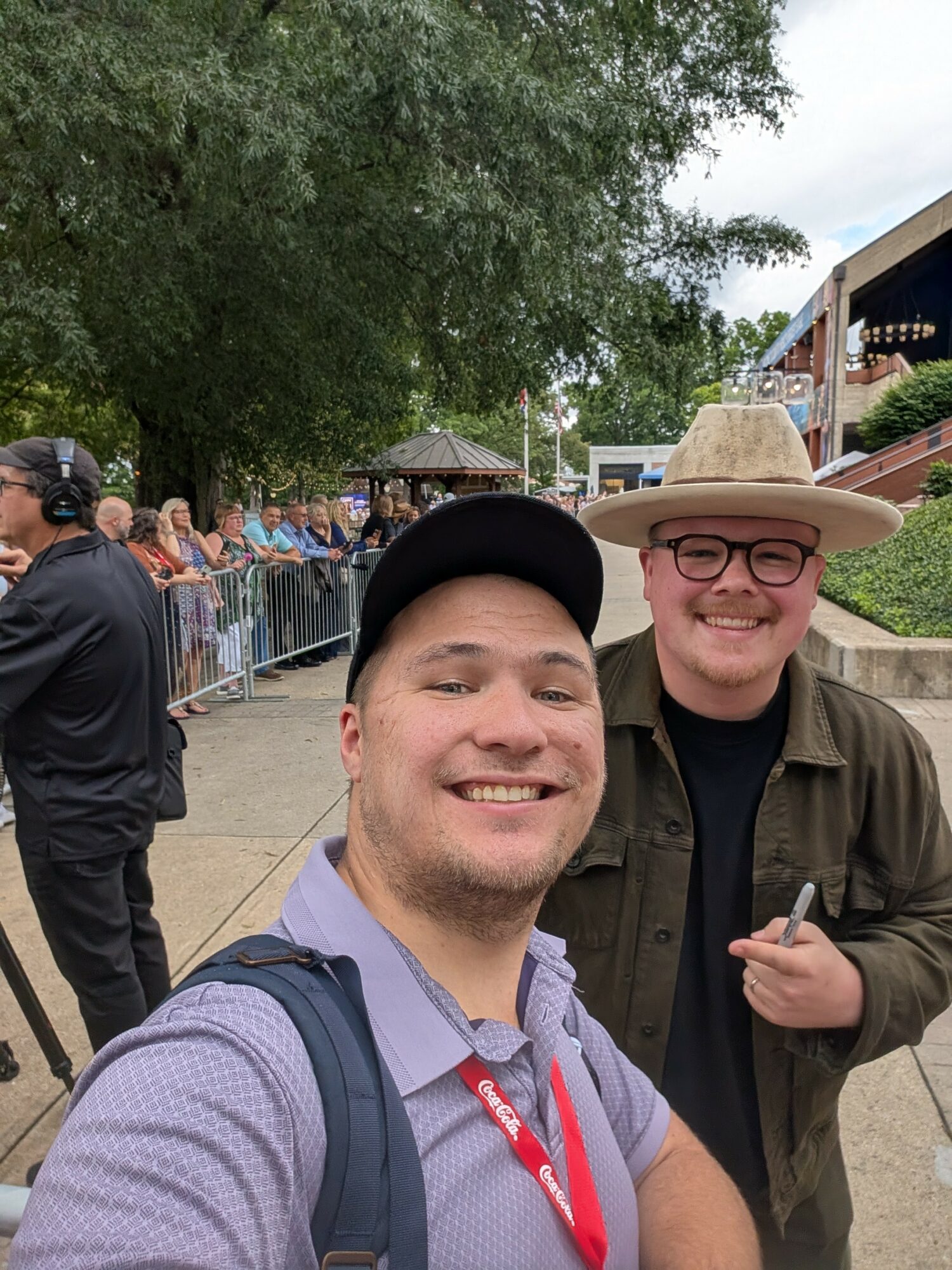 Two smiling men outdoors, one wearing a black cap and the other a straw hat, with trees and people in background.